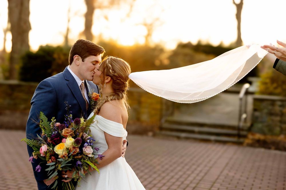 Newlywed couple shares romantic kiss under flowing veil during golden hour sunset at outdoor wedding ceremony.