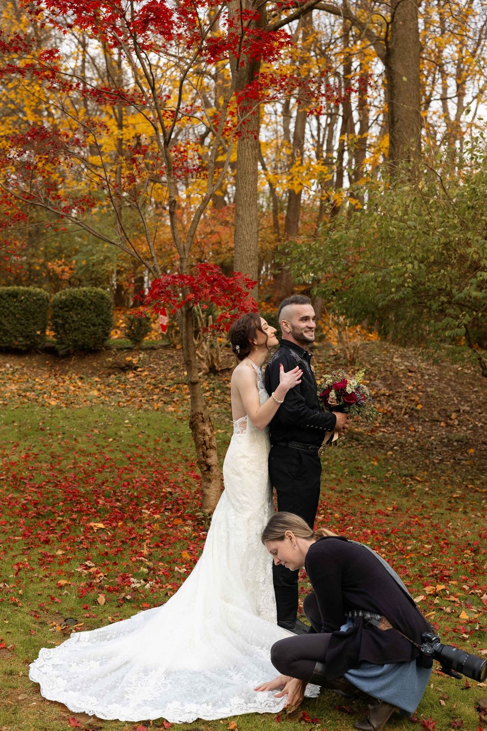 Wedding photographer takes photos of a couple in a scenic autumn forest setting with vibrant red and gold foliage.