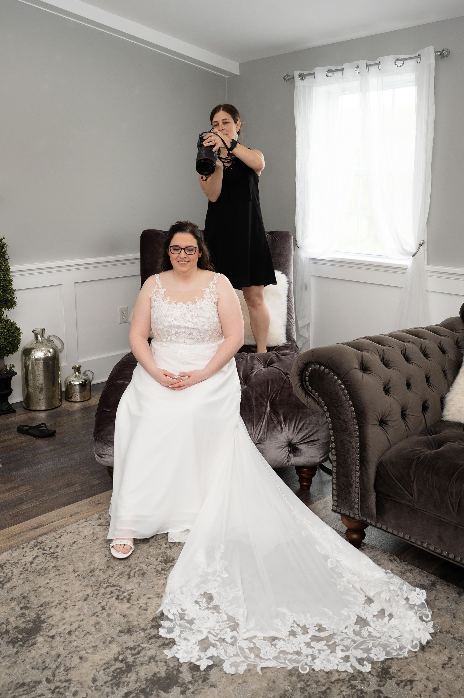 A bride in a white wedding dress sits elegantly on a gray tufted sofa while having her dress arranged.