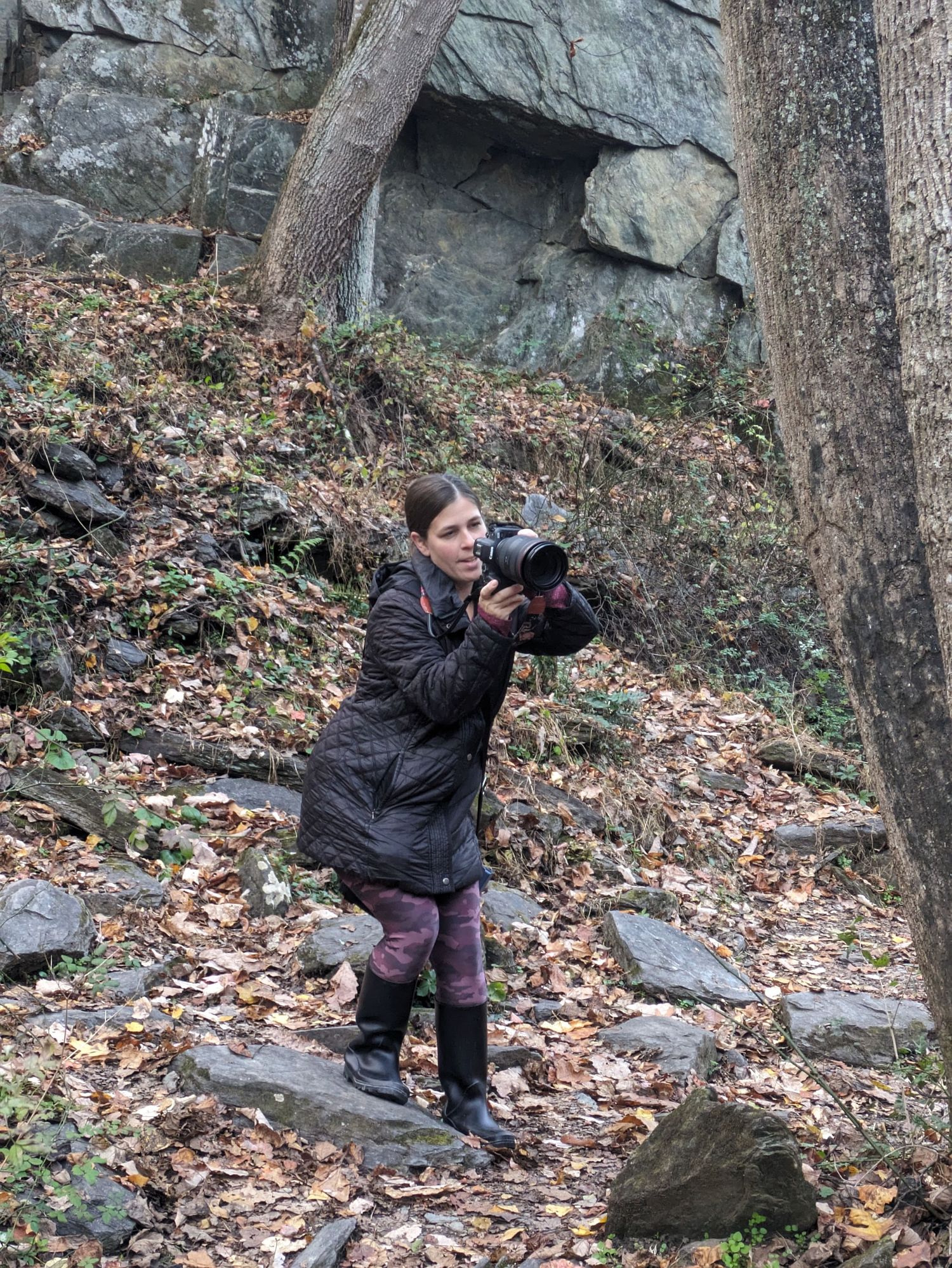 A photographer in a winter coat crouches on a rocky slope covered in fallen leaves while taking pictures in nature.