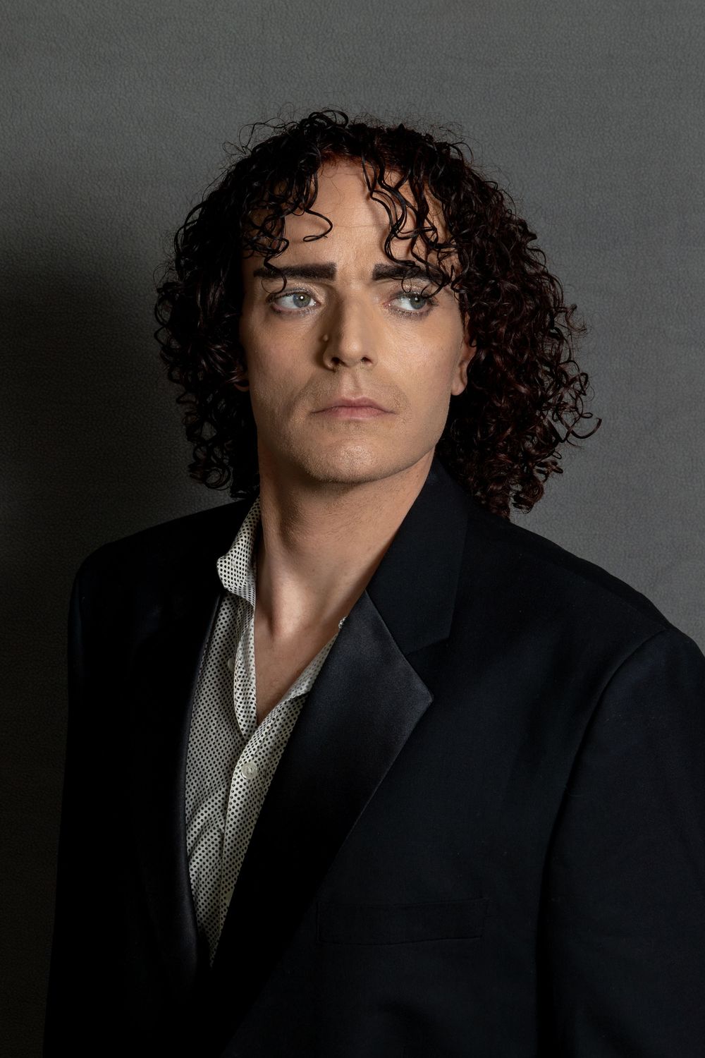 Studio portrait of a person with dark curly hair wearing a black blazer and light colored shirt against a dark background.