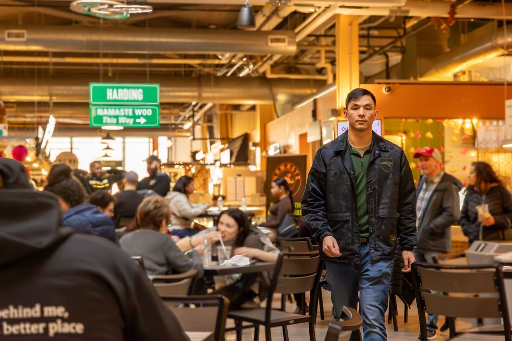 Person wearing a dark jacket stands in a busy food court with overhead directional signs and diners at tables.