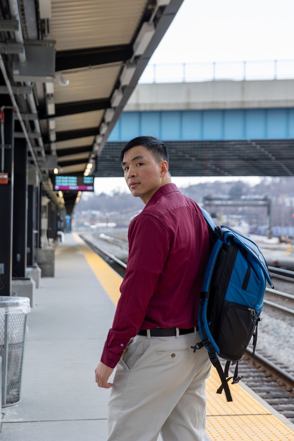 A person wearing a burgundy shirt and khaki pants waits on a train platform with a blue backpack.