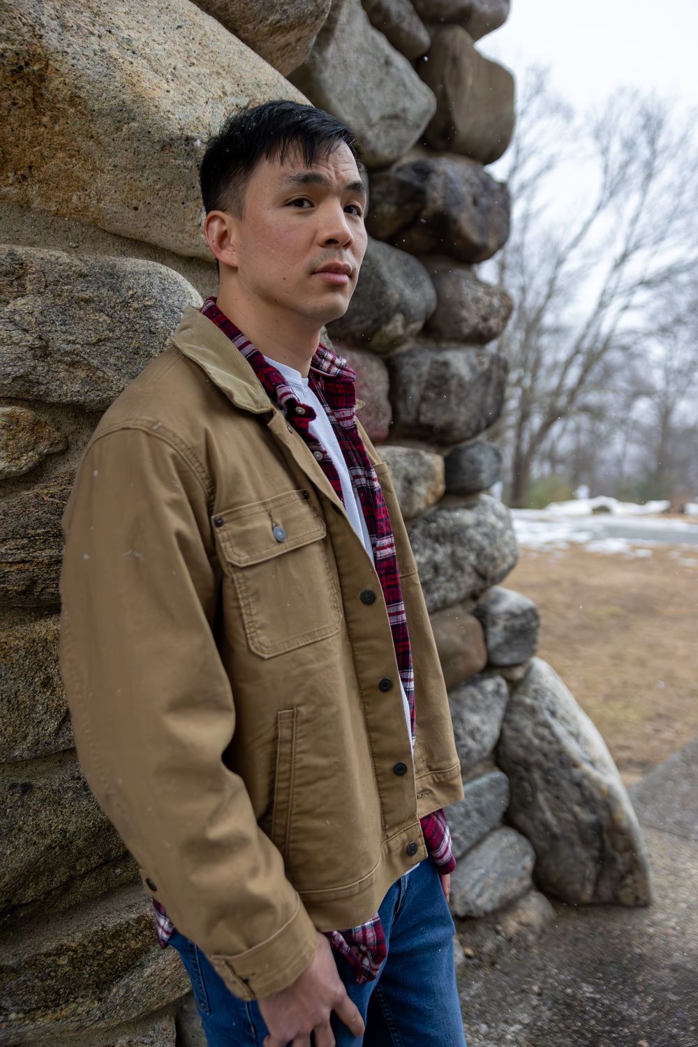 Person in tan work jacket and plaid shirt poses against stone wall in winter landscape.
