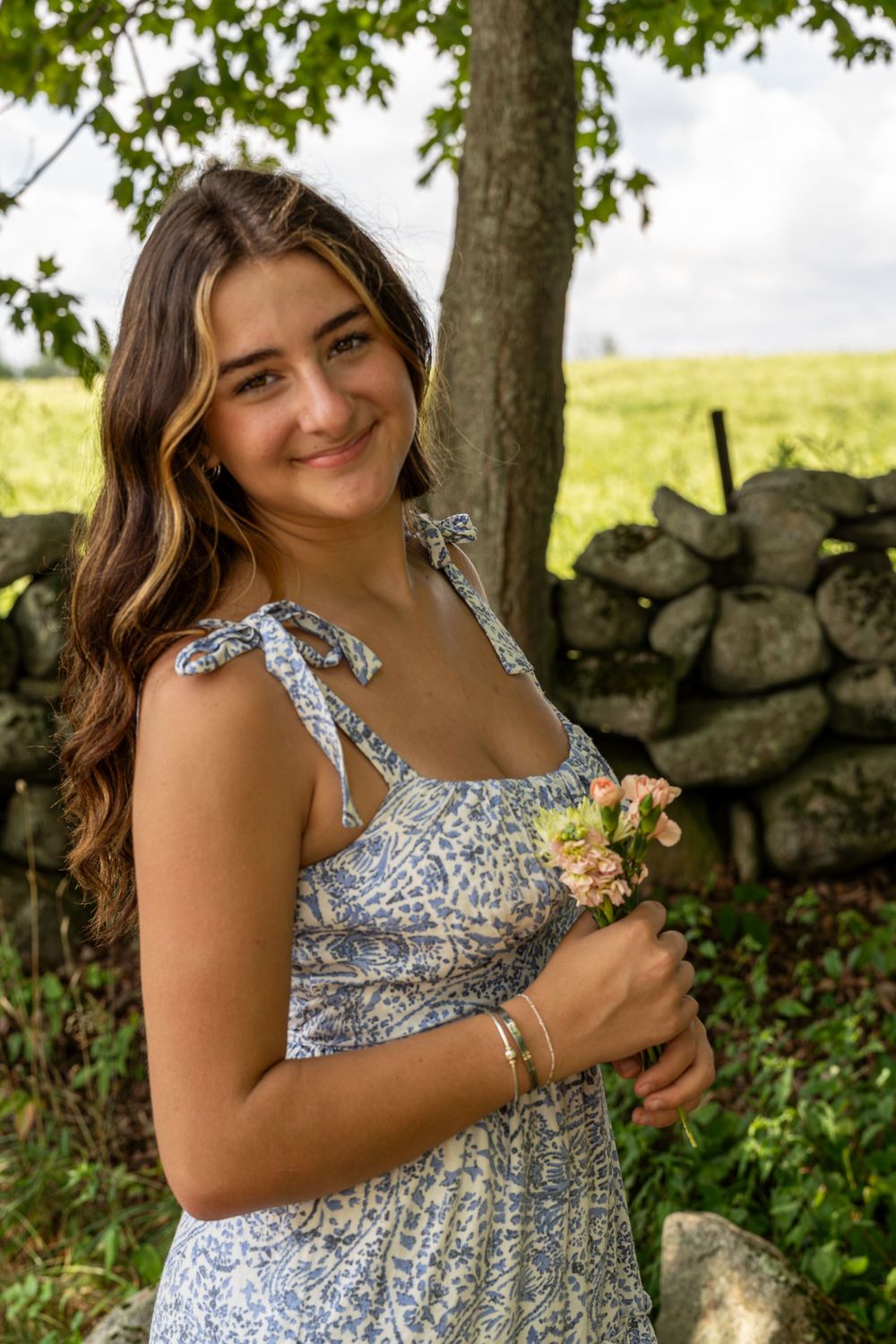 A person in a floral sundress poses outdoors against a stone wall and lush greenery on a sunny summer day.