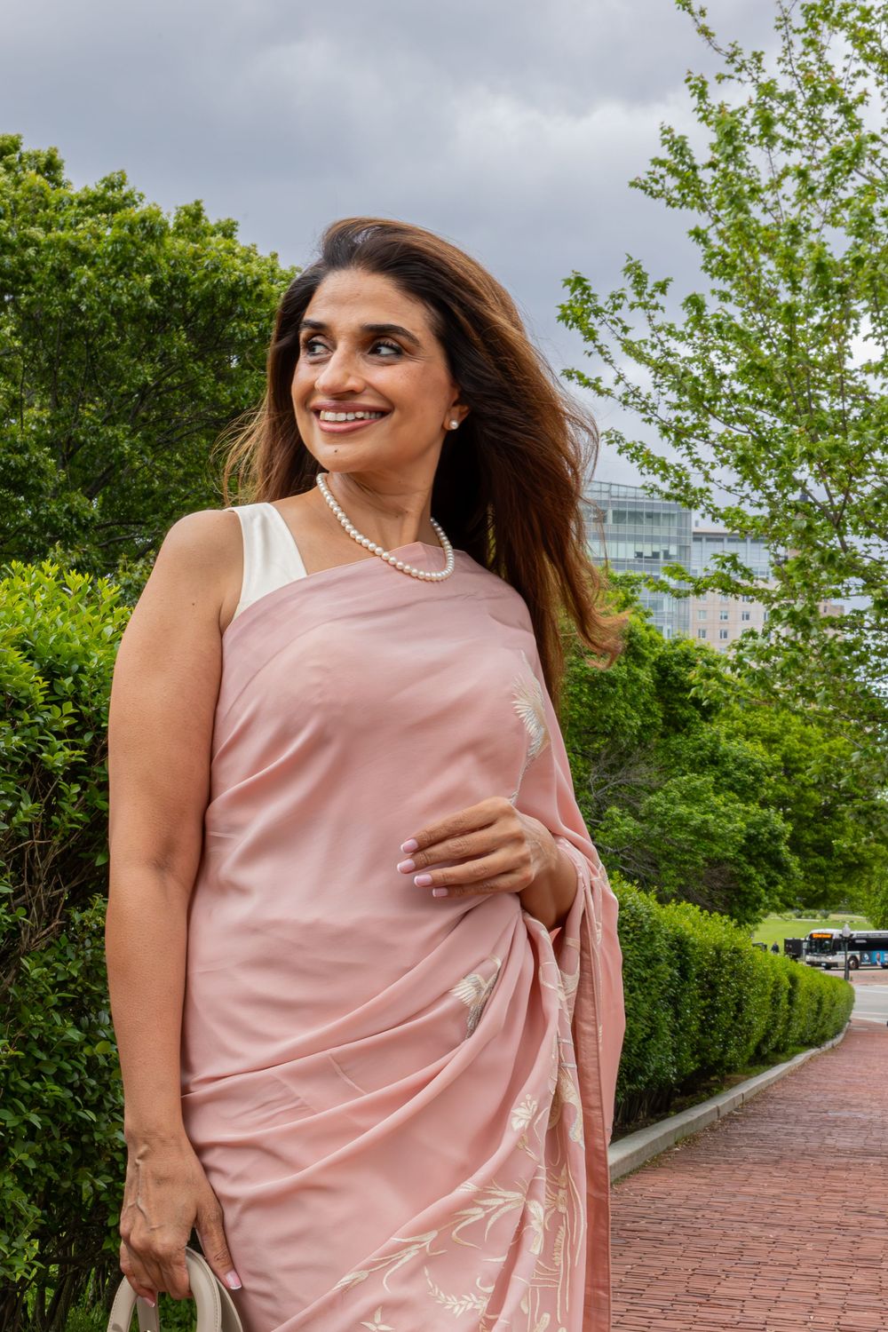A person in a blush pink sari and white blouse stands on a brick path surrounded by green foliage on a cloudy day.