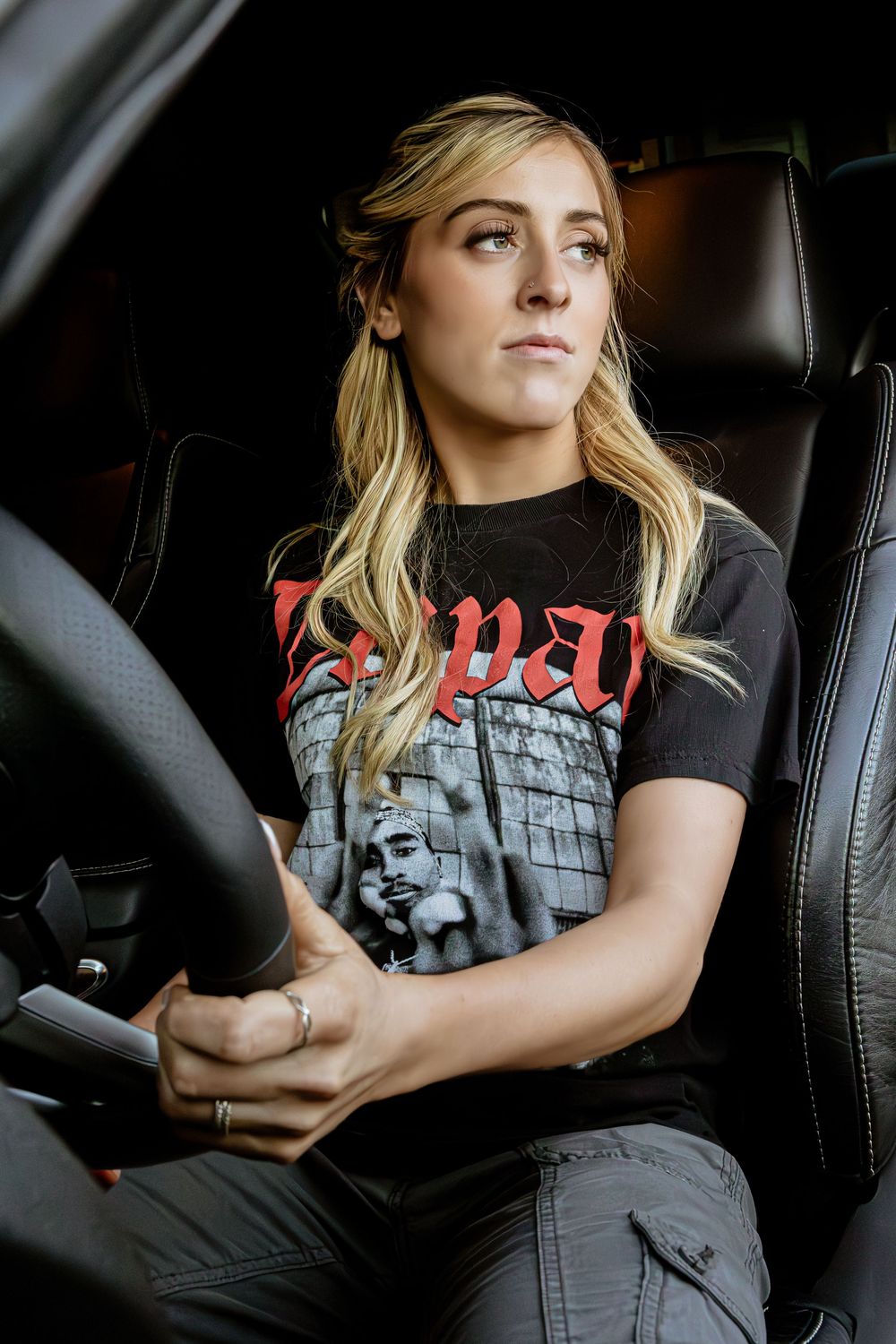 Person in black band t-shirt and cargo pants sits in driver's seat of car with blonde hair and rings on hand.
