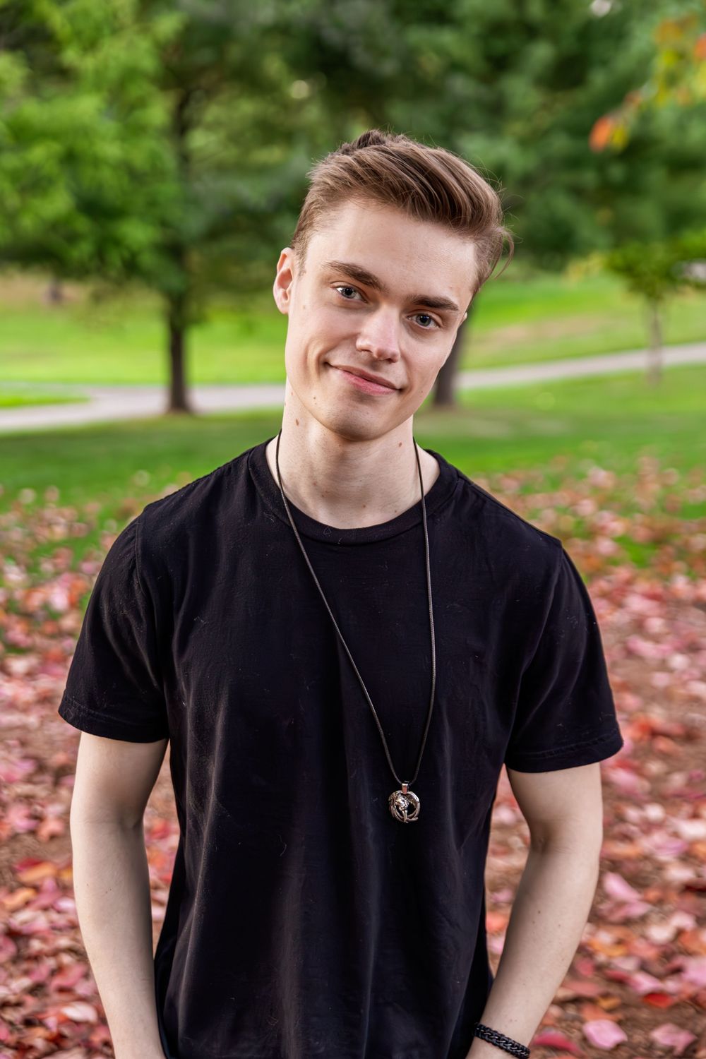 A person wearing a black t-shirt and necklace poses outdoors with autumn leaves scattered on the ground.