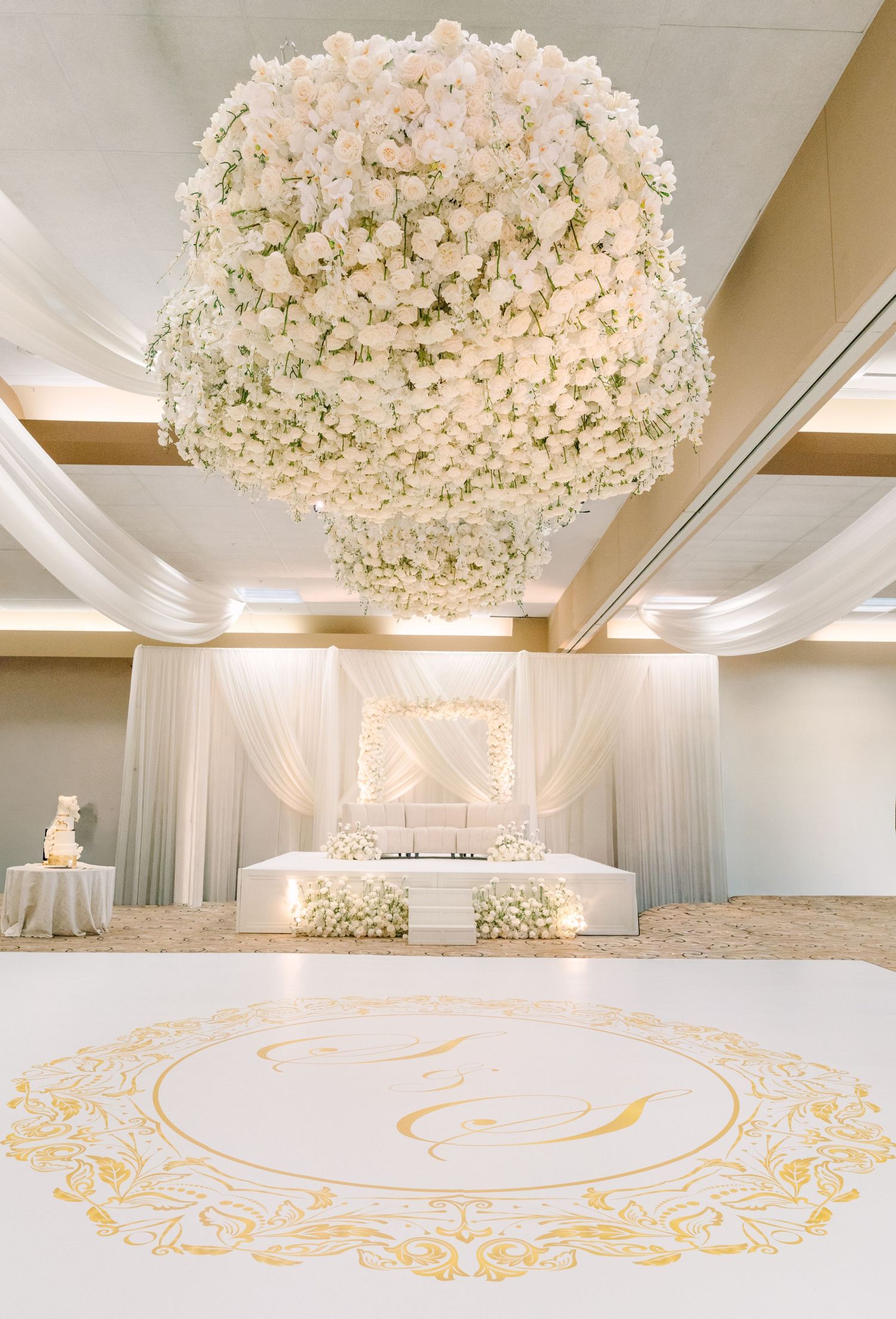 Elegant wedding ballroom with a large white floral chandelier, gold monogram dance floor, and draped white backdrop with sweetheart table.