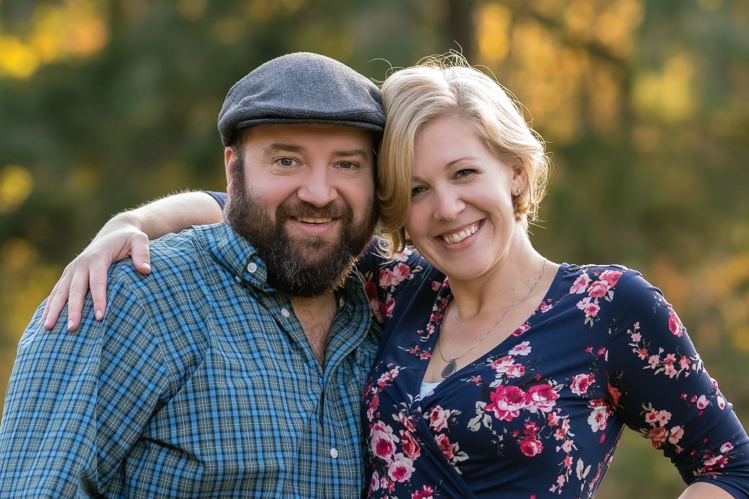 A smiling couple poses outdoors in autumn, man in plaid shirt and cap, woman in floral dress, embracing warmly.