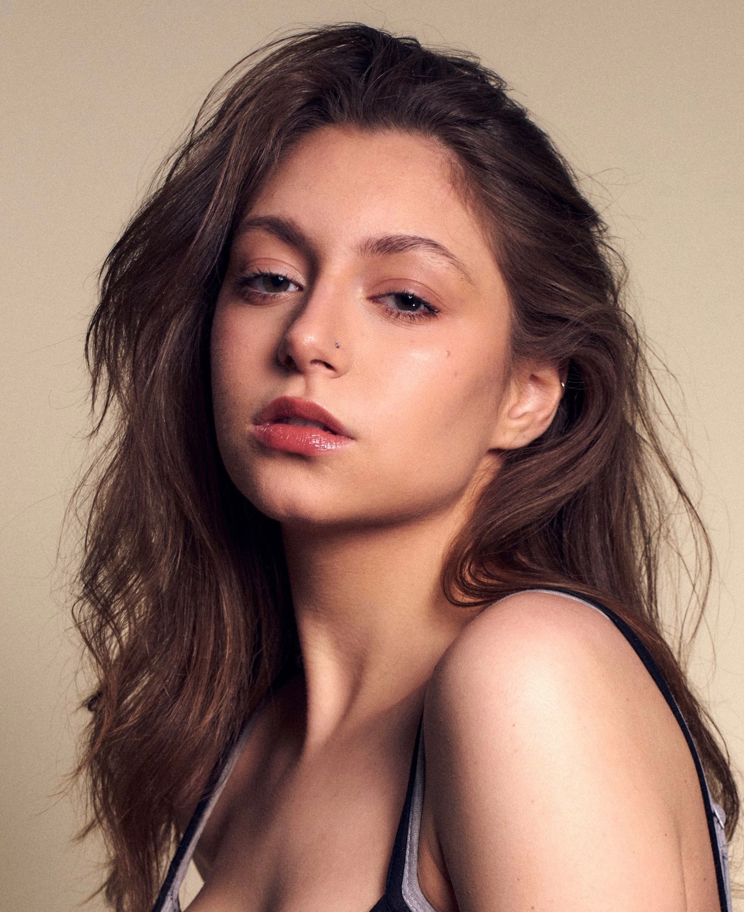 Young woman with long wavy brown hair and natural makeup posing against a warm beige background in a sleeveless top.
