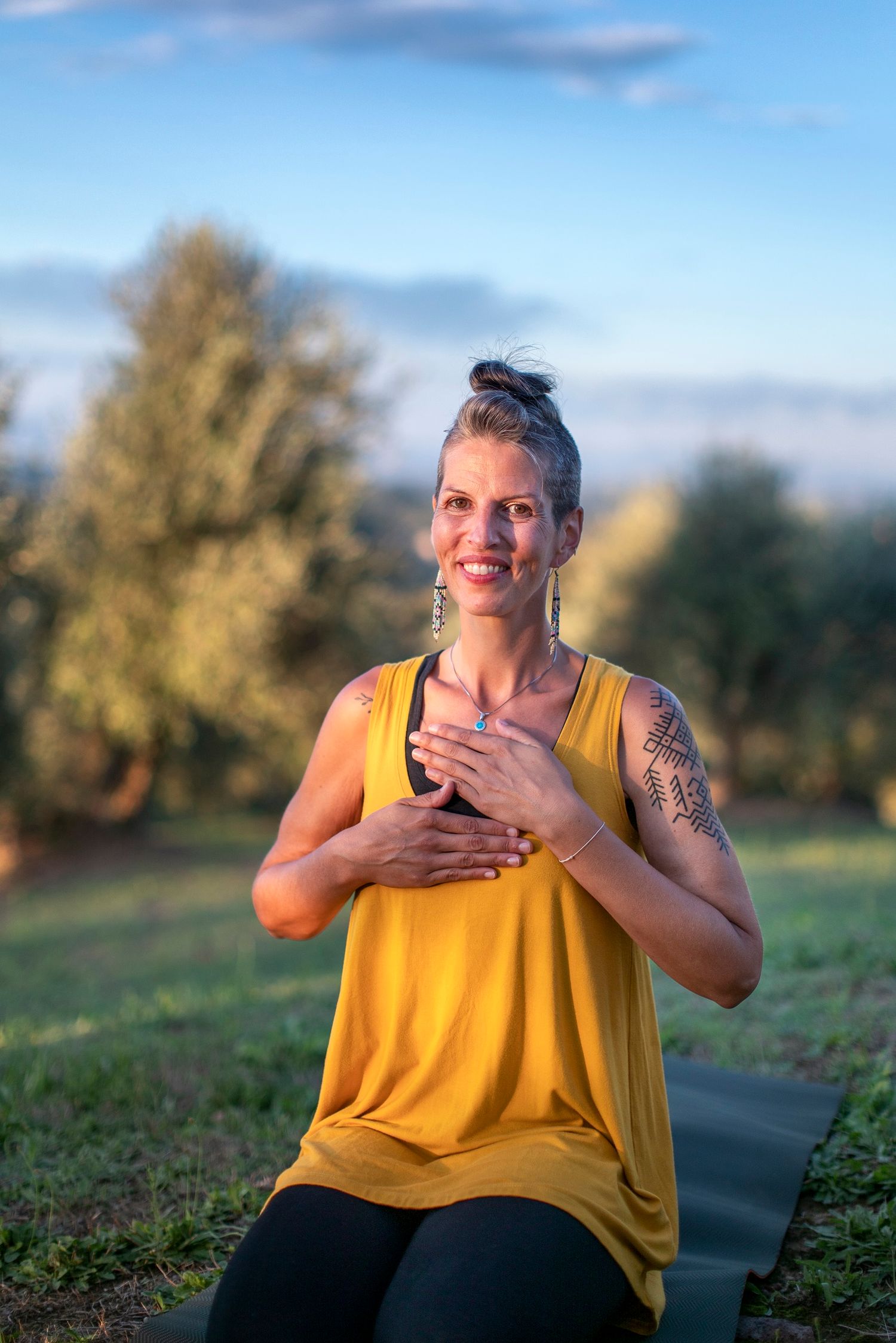 Smiling woman in yellow tank top with tattoo sitting on yoga mat in sunny outdoor field.