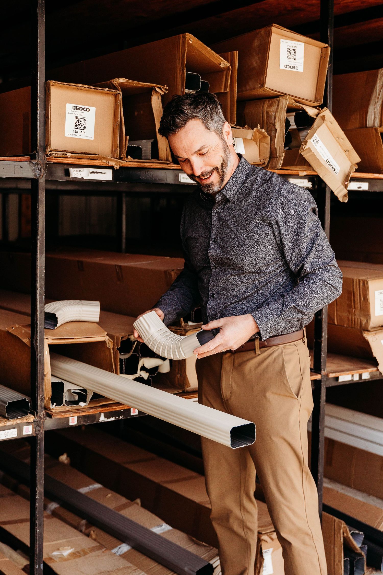 Warehouse worker in gray pullover and khakis examines inventory on industrial shelving with cardboard boxes.