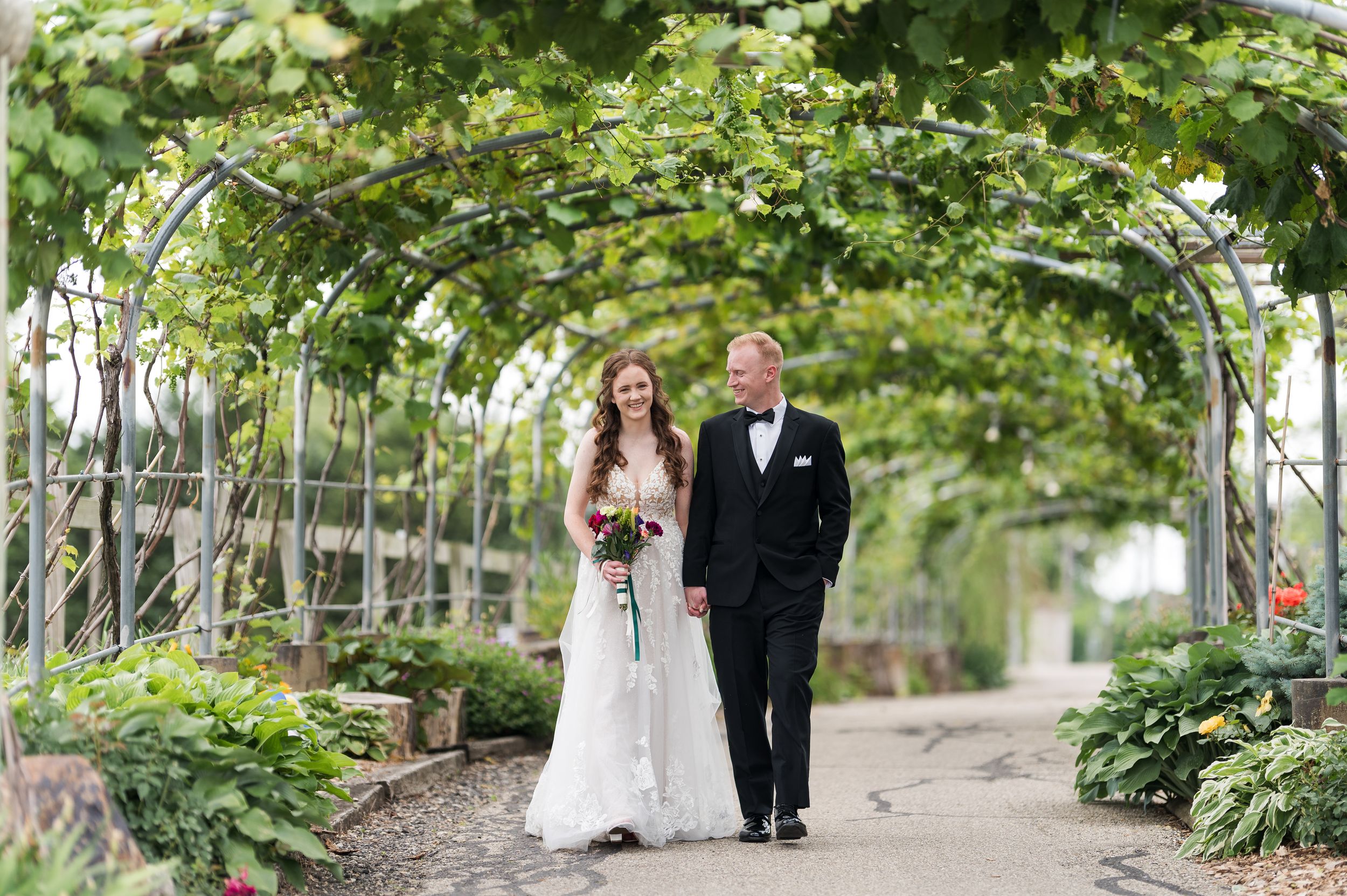 A couple walks down a romantic garden path lined with lush greenery and overhead vines during a wedding ceremony.