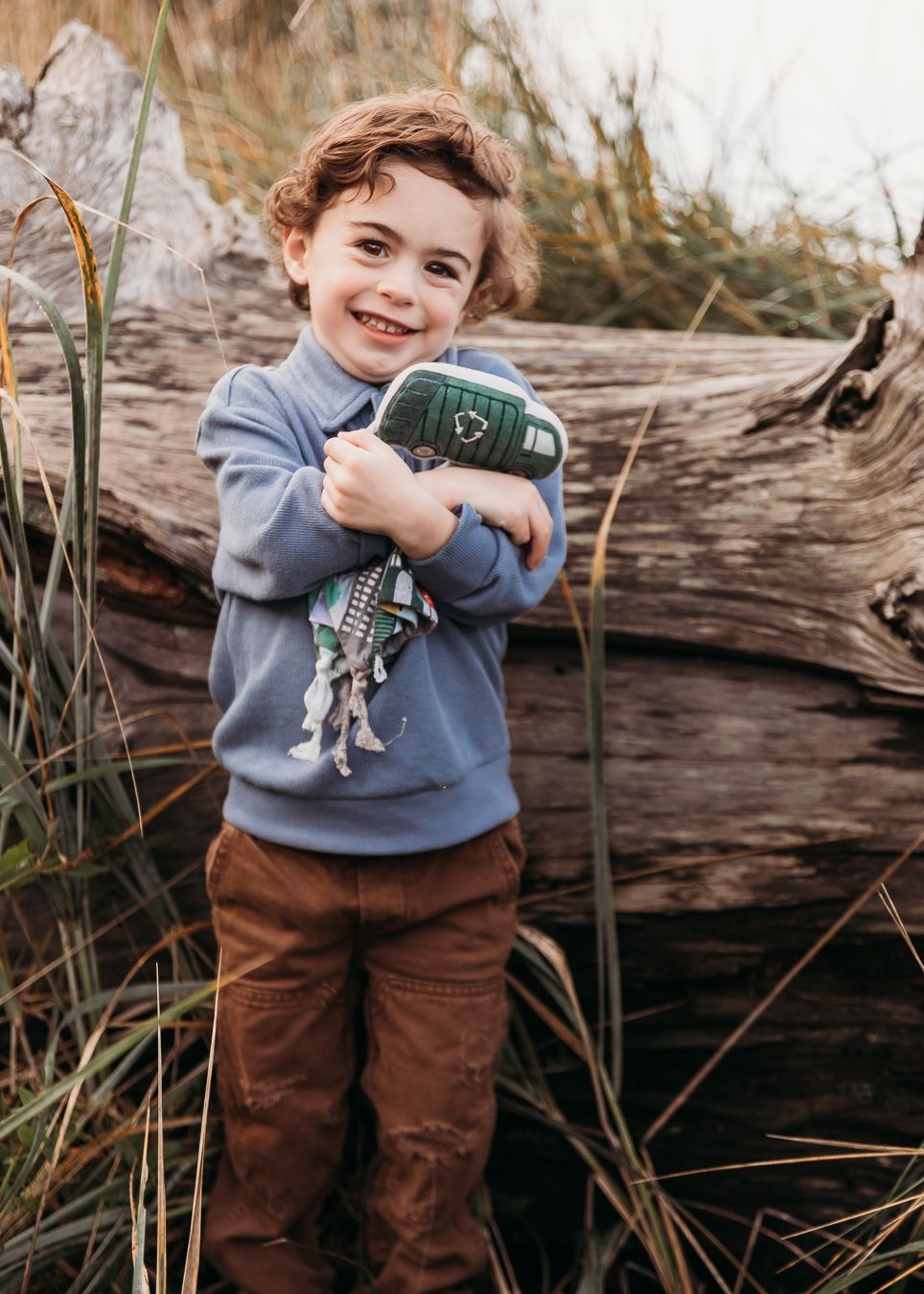 A child in a blue sweater and brown pants plays outdoors near a fallen log and tall grass in a natural setting.