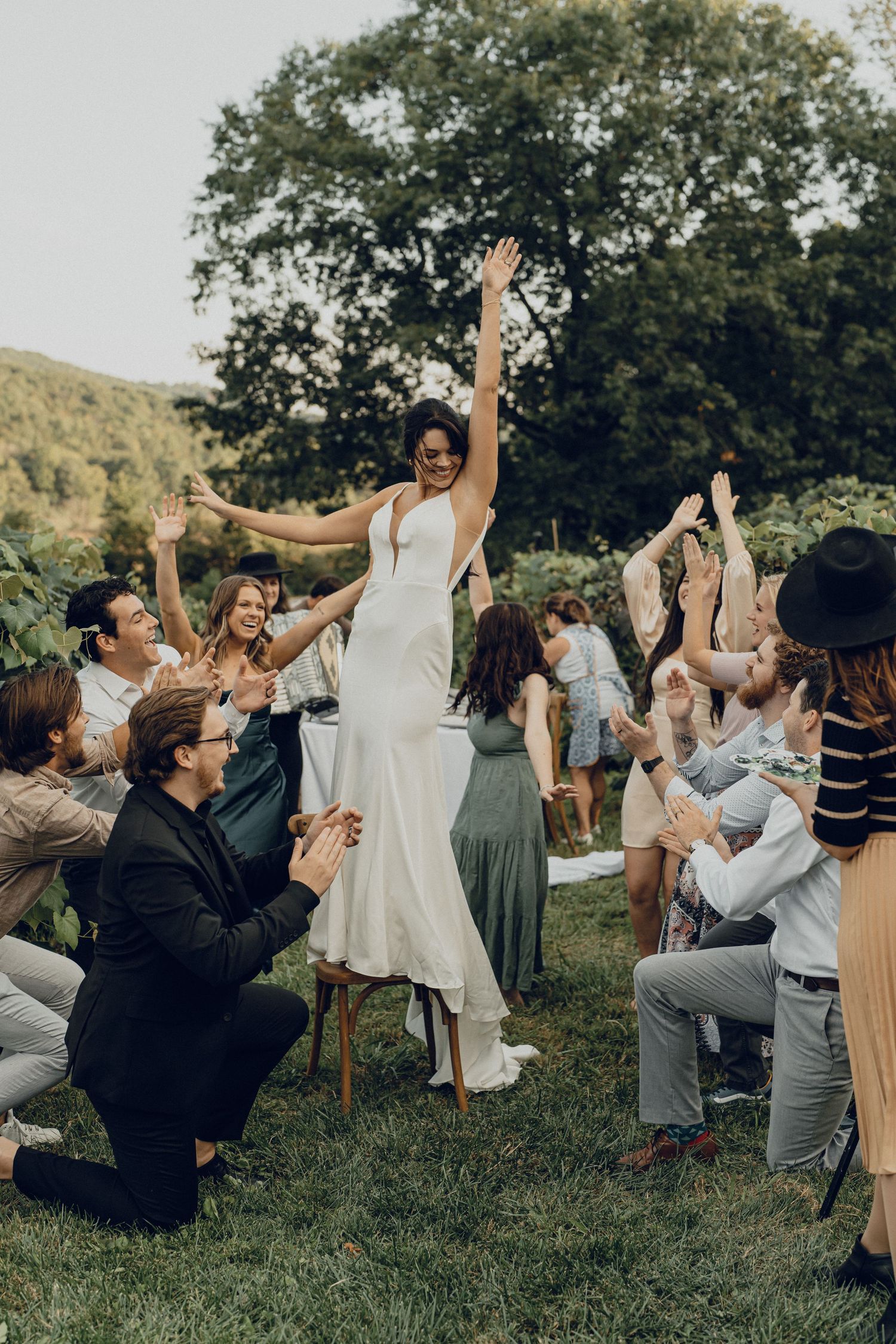 Bride in white gown stands on chair with arms raised, celebrated by cheering wedding guests outdoors.