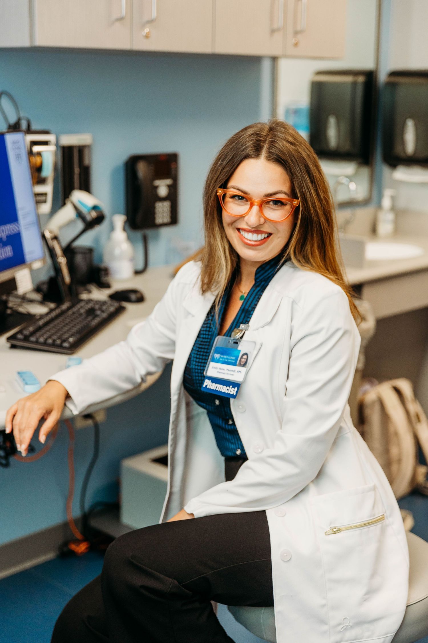 Medical professional in white lab coat and glasses works at computer workstation in clinical laboratory setting.