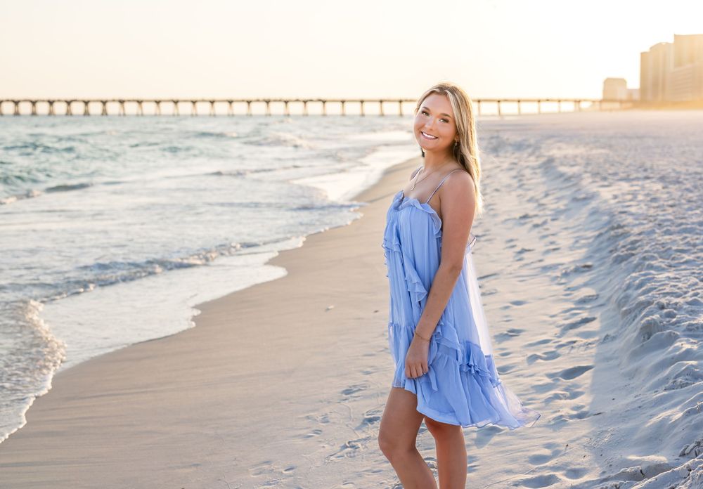 Young woman in blue dress smiling on sandy beach at golden hour with pier in background.