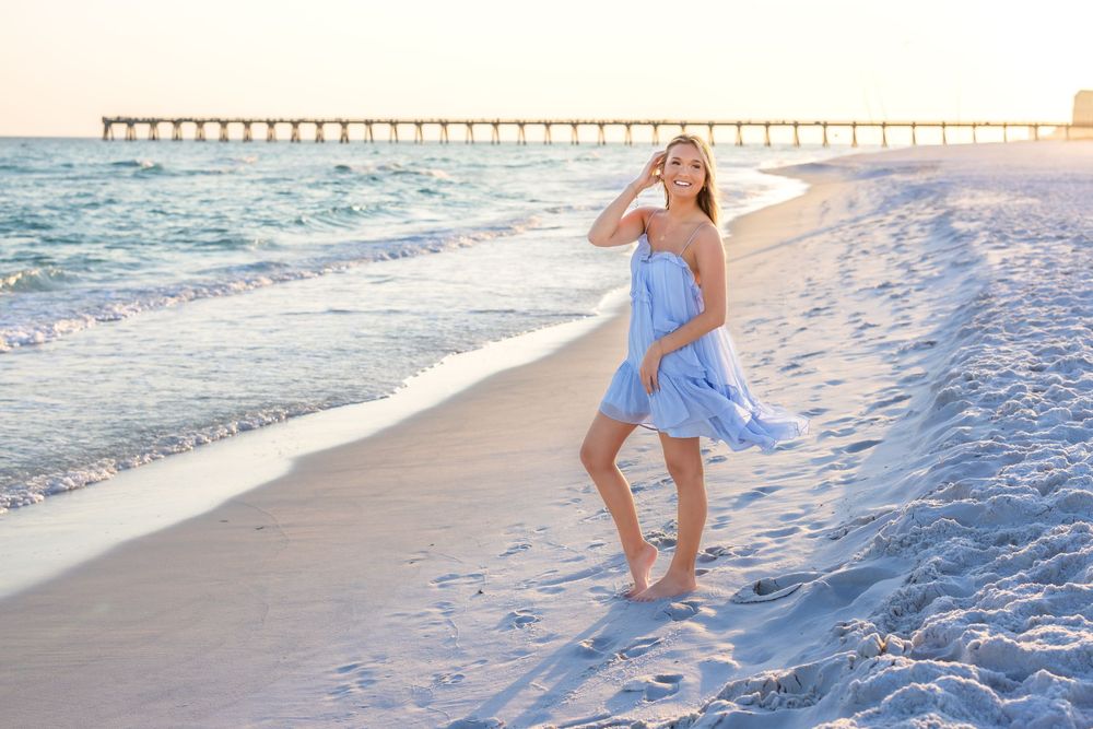 A young woman in a flowing blue dress walks barefoot on a sandy beach at sunset with a pier in the background.