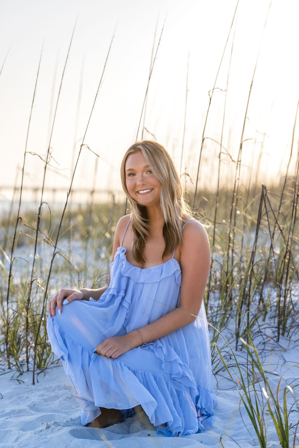 Young woman in blue dress posing on sandy beach dunes with sea oats at golden hour sunset.