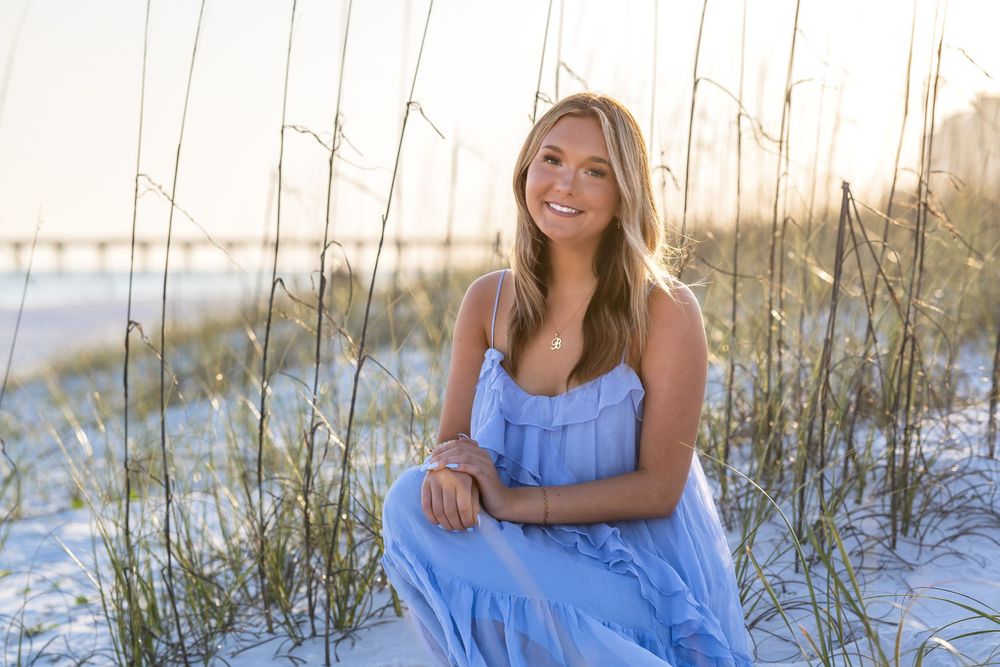Young blonde woman in blue dress sitting on white sand beach dunes with sea oats and pier at sunset.