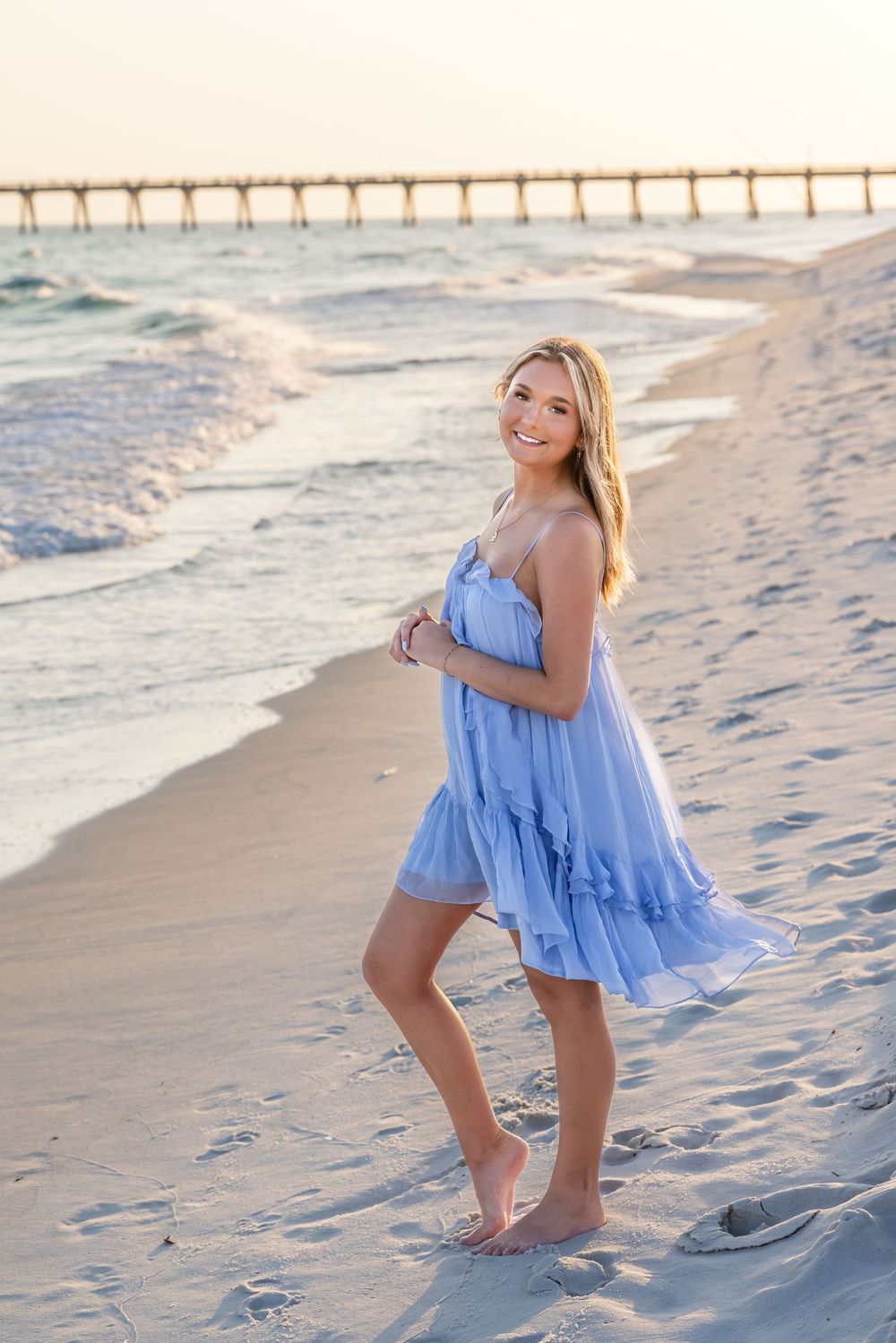 Smiling blonde woman in a light blue sundress standing barefoot on a sandy beach at sunset with a pier in the background.