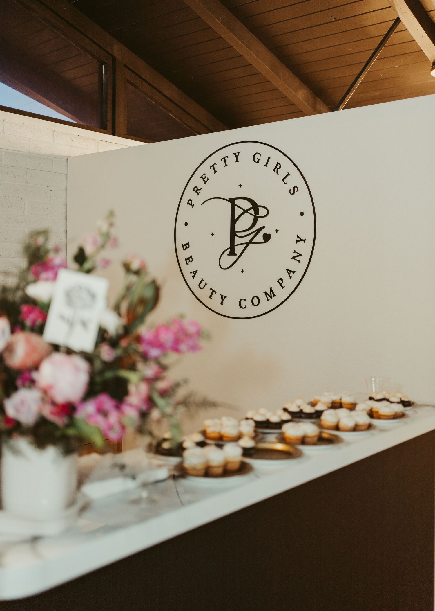 Pretty Girls Beauty Company branded dessert table with cupcakes and pink floral arrangement at an elegant event.