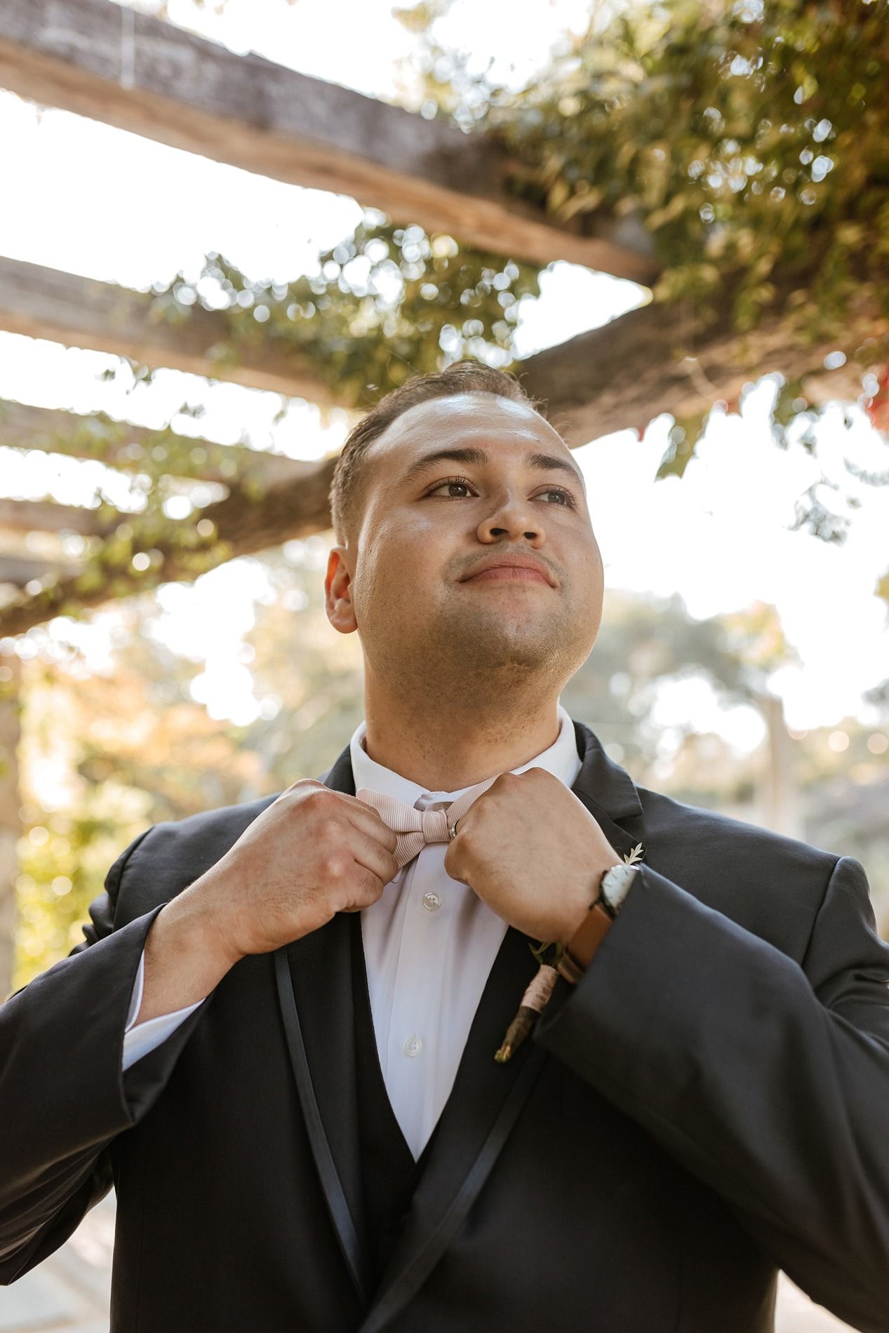Groom in black tuxedo adjusting bow tie outdoors under wooden pergola with greenery on wedding day.