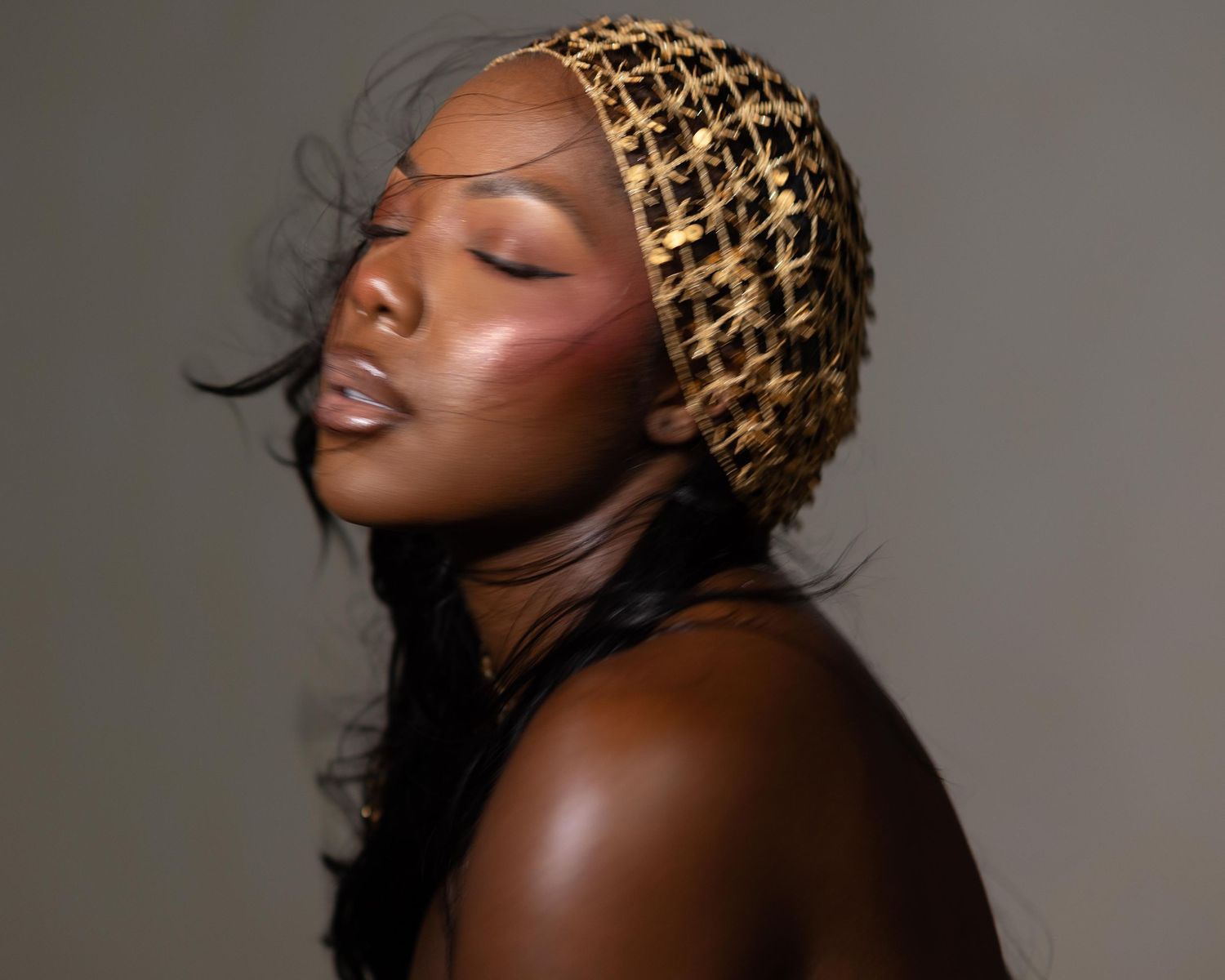 A woman with dark skin wears a gold and black net headscarf, eyes closed, with rosy blush makeup.