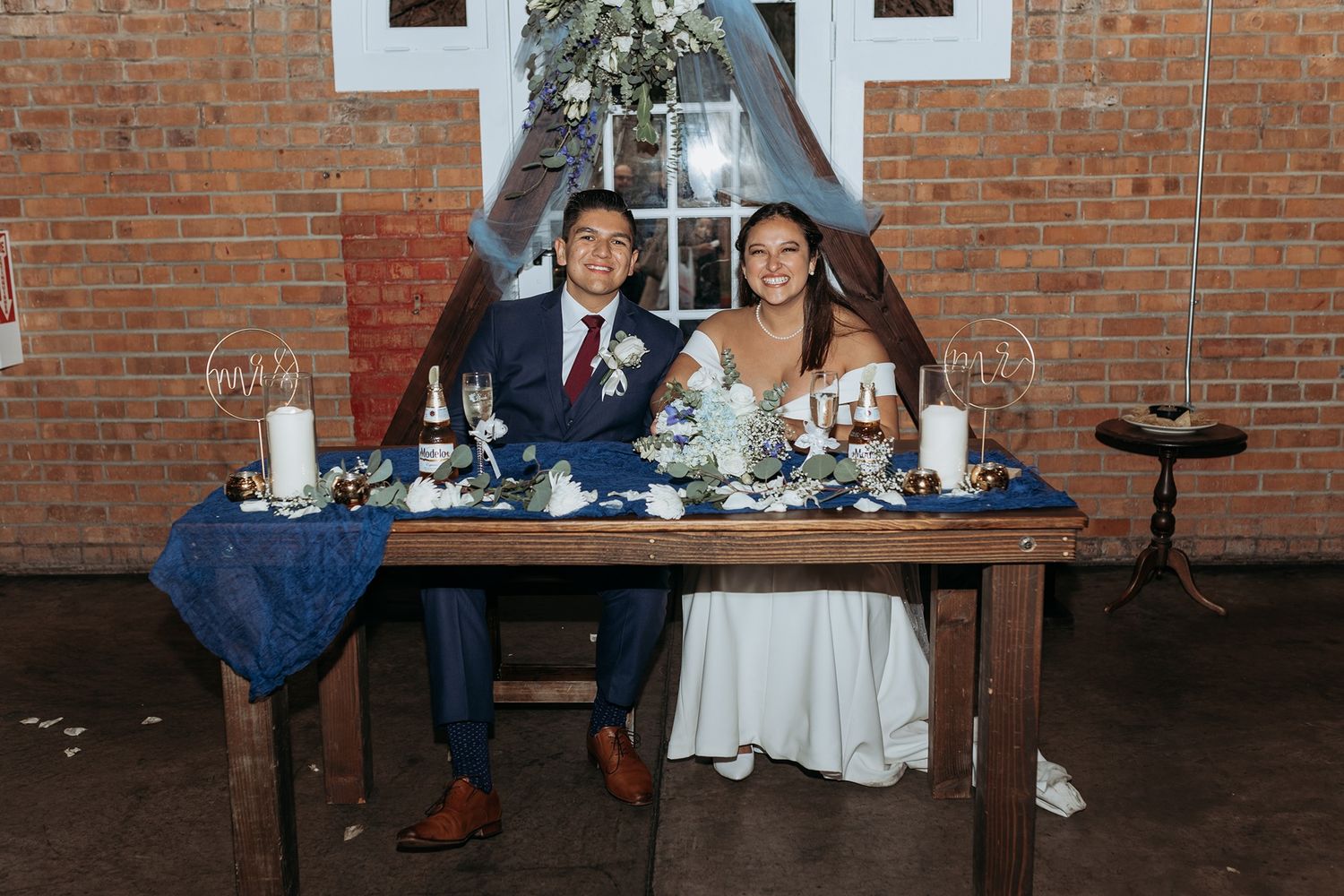 A couple sits together at a rustic reception table with blue decor against an exposed brick wall.