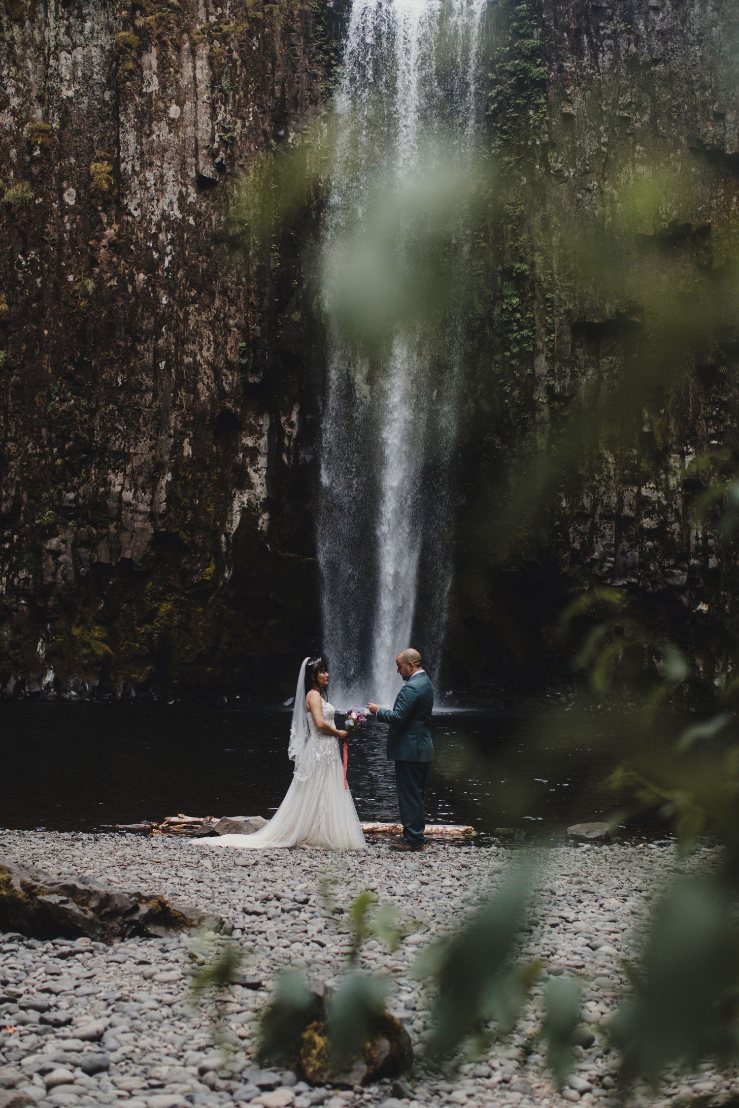 A couple shares an intimate moment by a majestic waterfall in a lush forest setting.