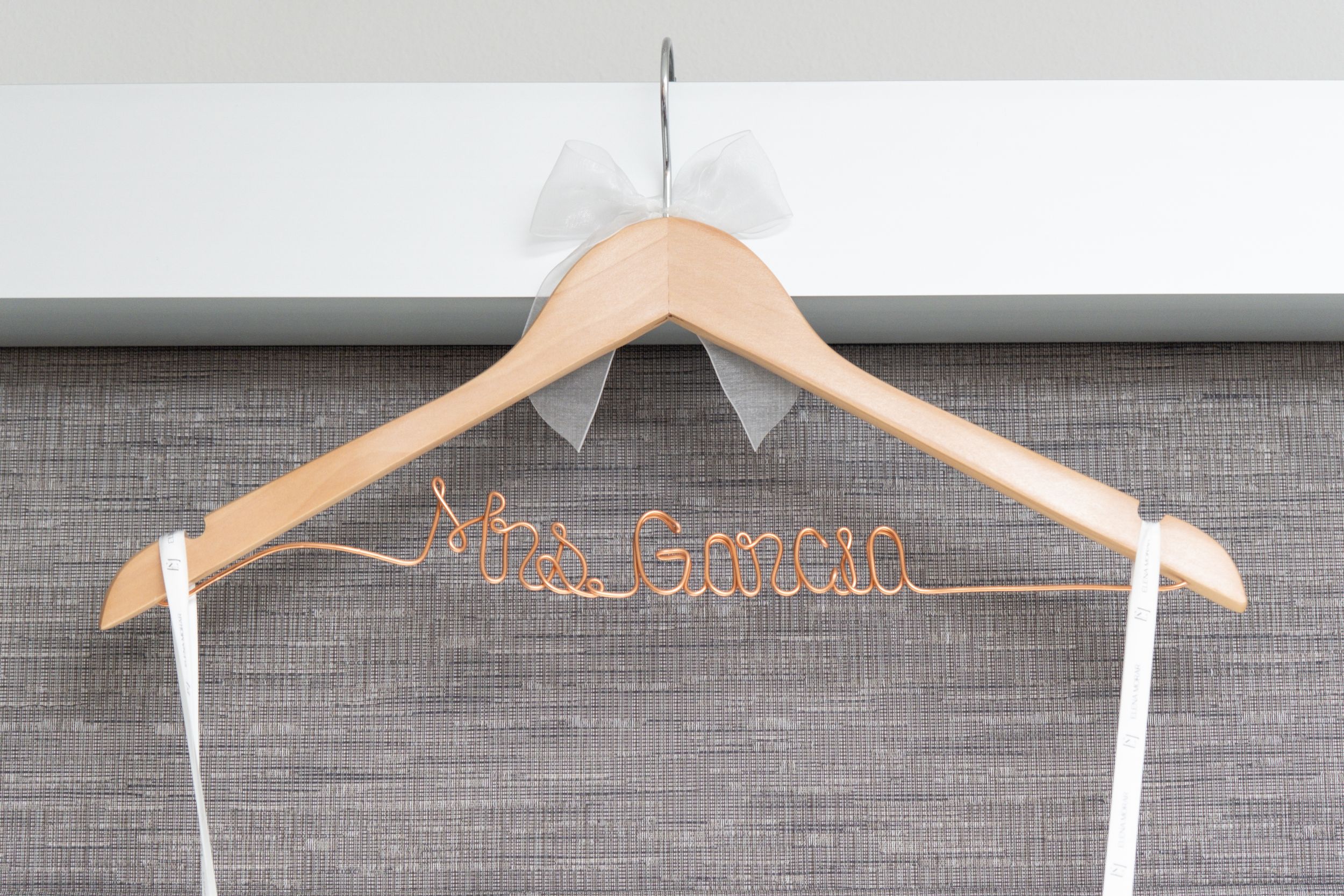 A wooden wedding hanger with 'Mrs. Barrera' written in rose gold wire against a gray textured background.