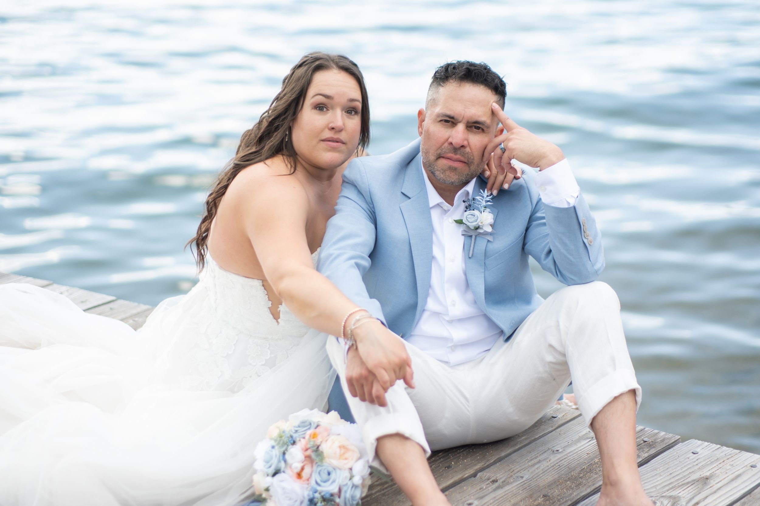 A couple in wedding attire sits together on a dock by the water on their special day.