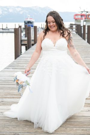 A joyful moment captured on a wooden dock as someone in a flowing white wedding gown holds their dress while smiling.