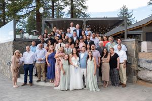 Large wedding party group poses together outdoors at venue with pine trees in background.