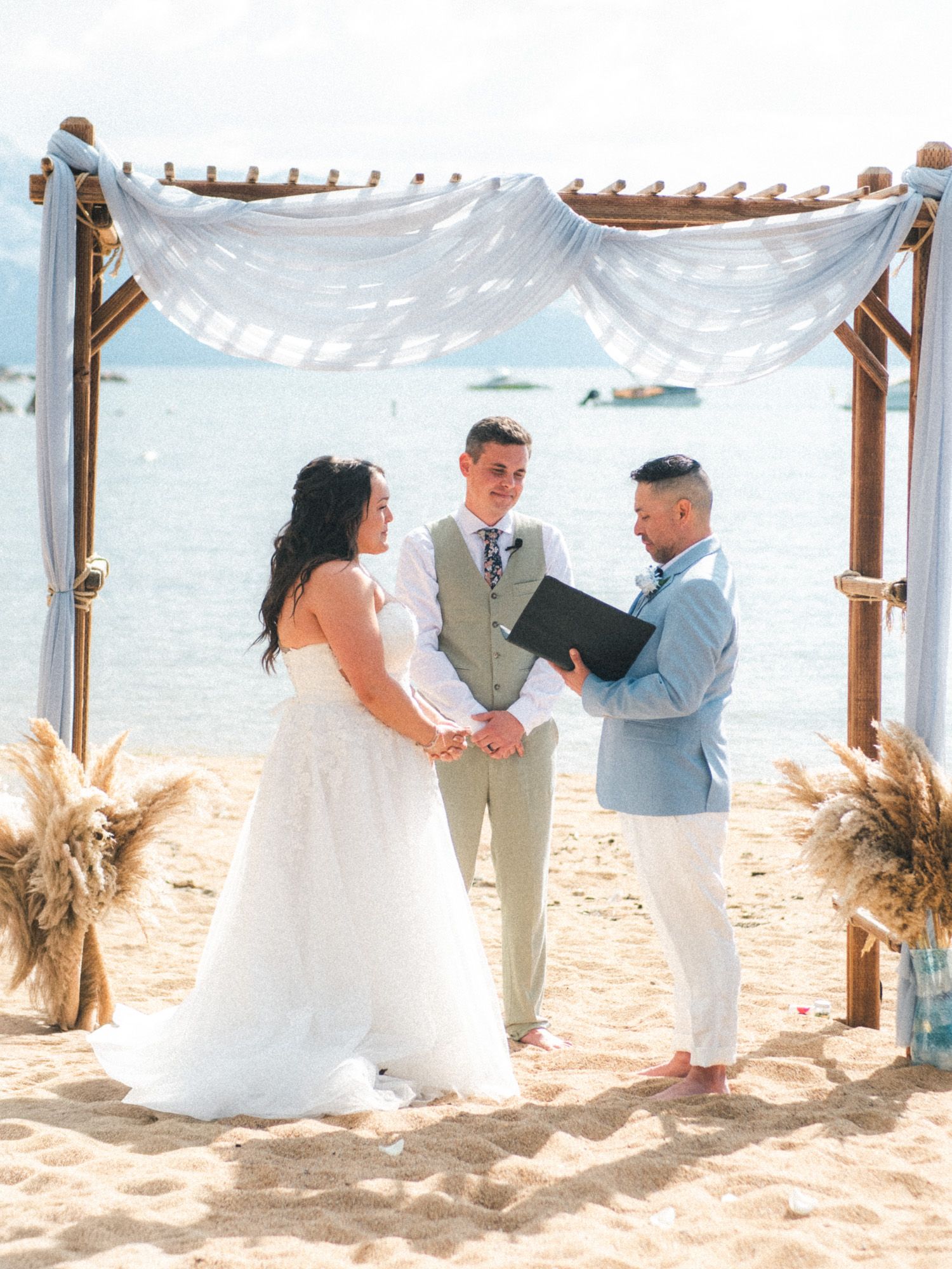 A beachside wedding ceremony under a white draped pergola decorated with pampas grass on a sunny tropical day.