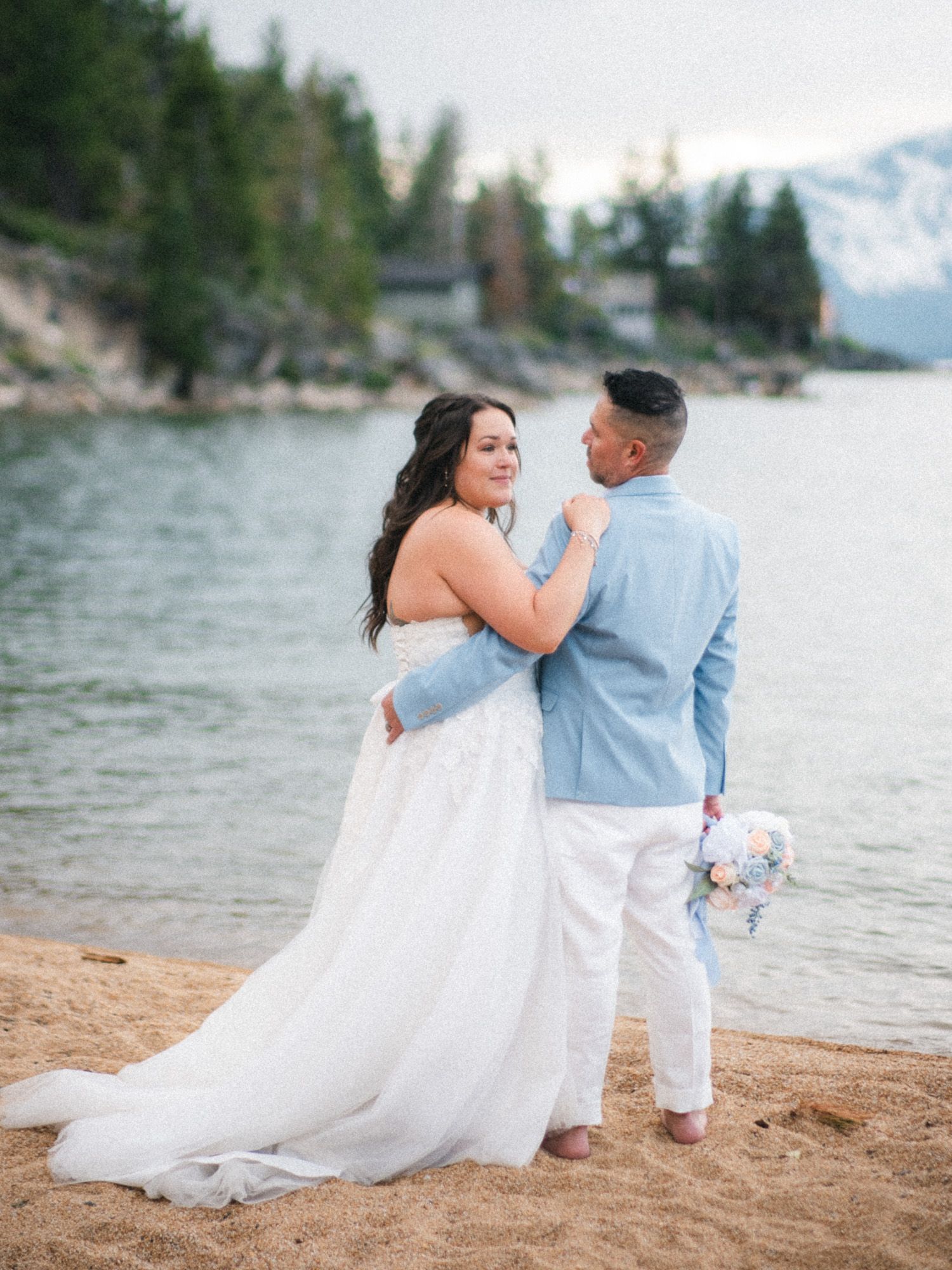 A couple sits together on a beach by a lake with mountains and pine trees in the background.
