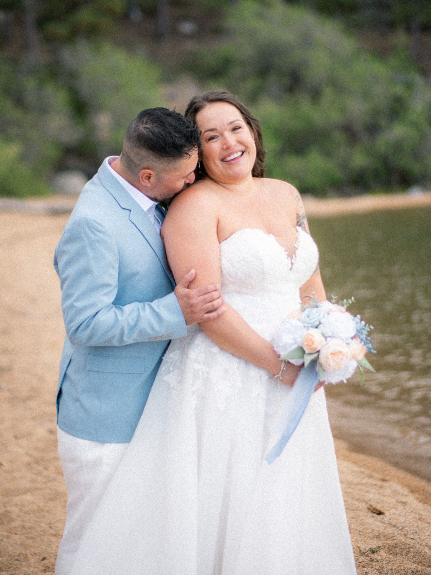 A romantic wedding couple embraces by a lakeside wearing light blue suit and white gown during an outdoor photo session.