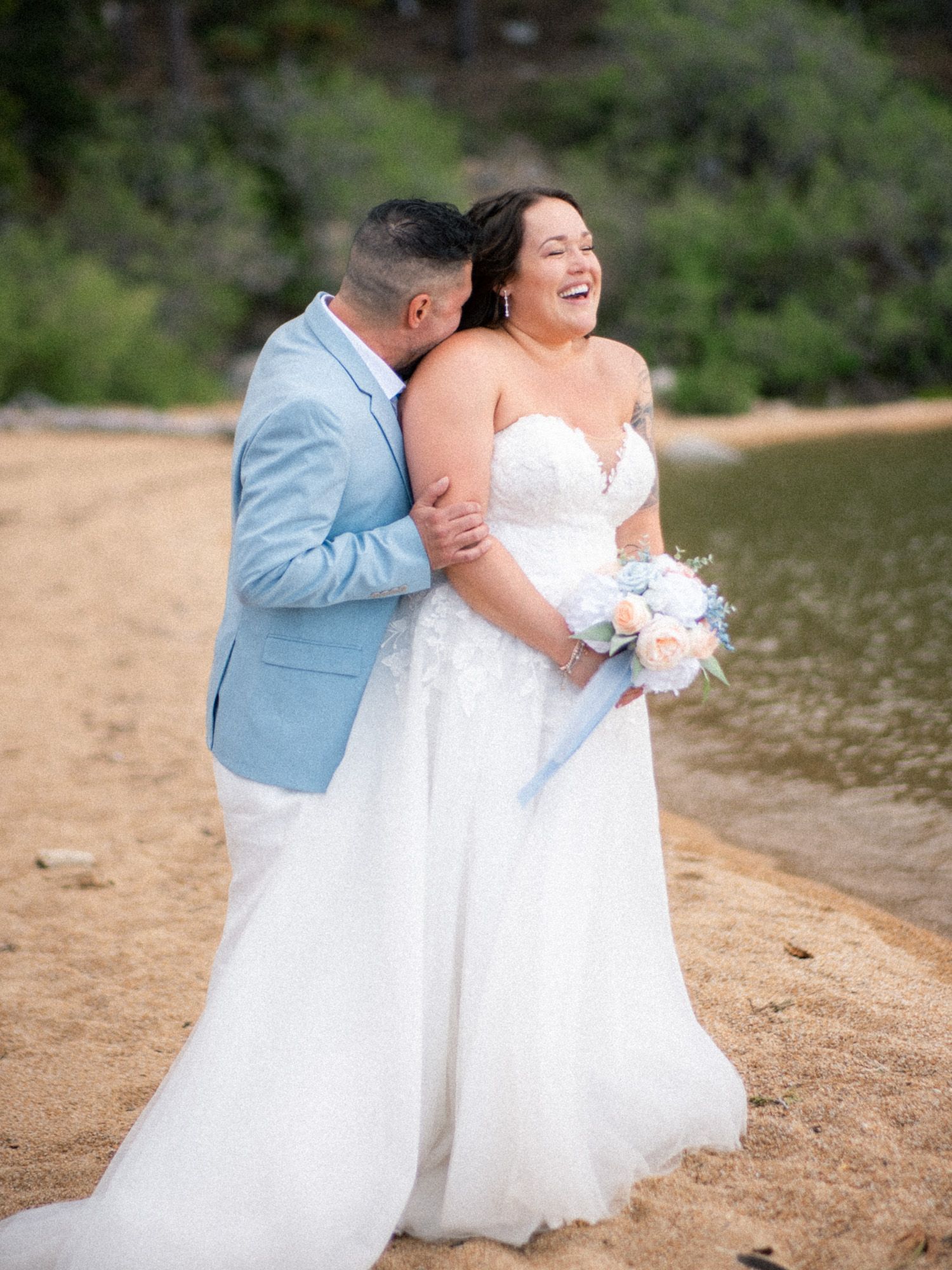 A couple in wedding attire shares a tender moment on a sandy beach with trees in the background.