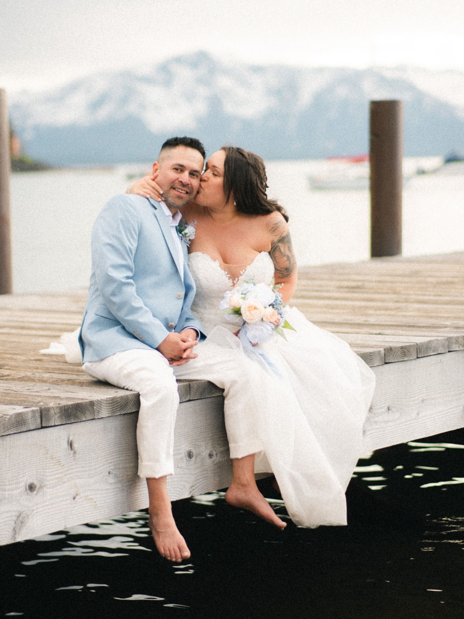 Romantic couple shares intimate moments on a wooden dock by a lake with mountain views in the background.