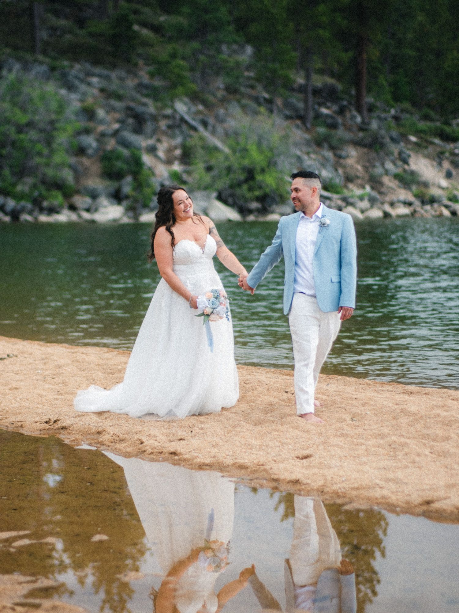 A couple in wedding attire walks along a sandy beach shore with reflections in the calm lake water and forest backdrop.