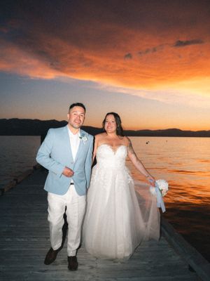 Wedding couple poses at sunset on a dock by Lake Tahoe with dramatic orange and pink sky reflecting on calm waters.