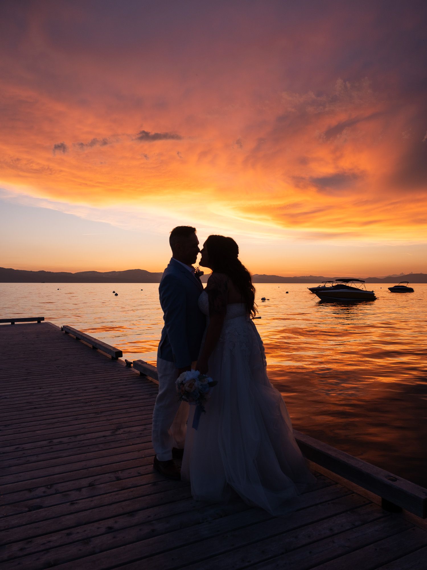 Silhouetted couple shares romantic moments at sunset on a wooden dock by the lake with dramatic orange sky.
