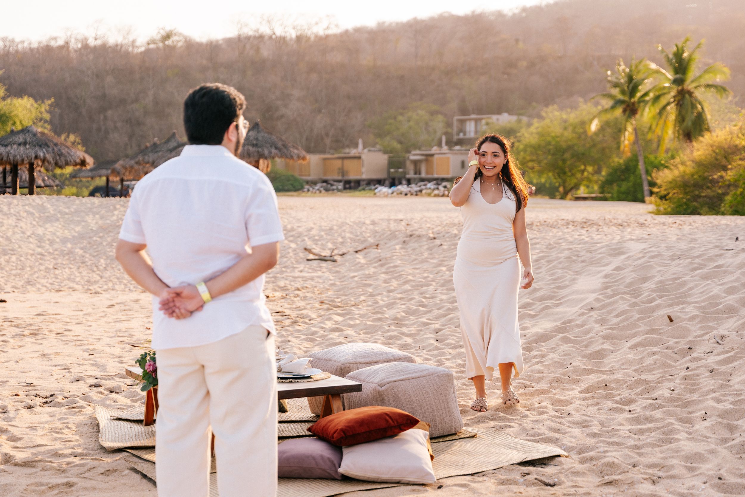 A romantic beach picnic setup at sunset with white cushions, blankets and decorative elements on the sand.
