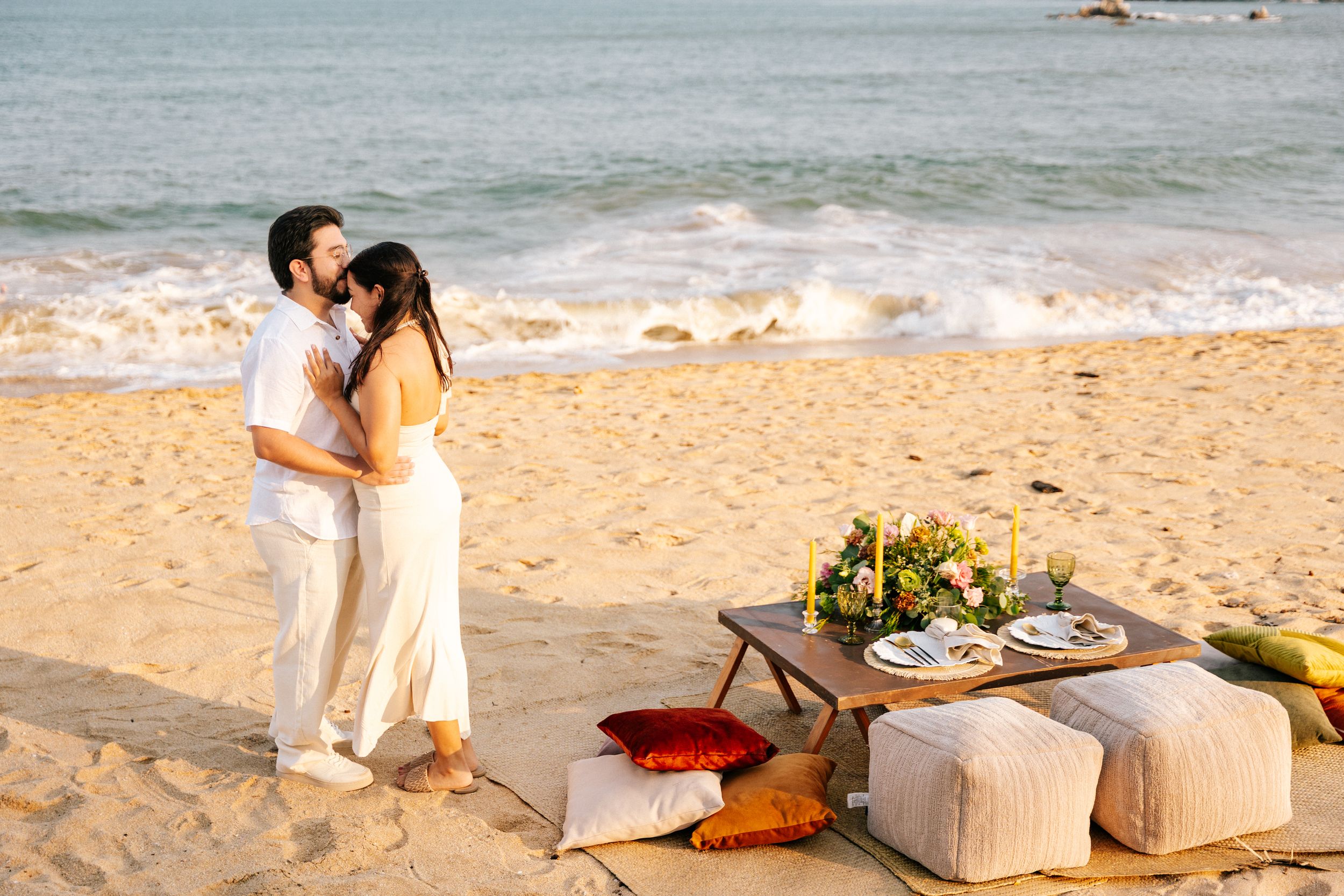 A romantic beach wedding setup with white decor and floral arrangements along a sandy tropical shoreline at sunset.