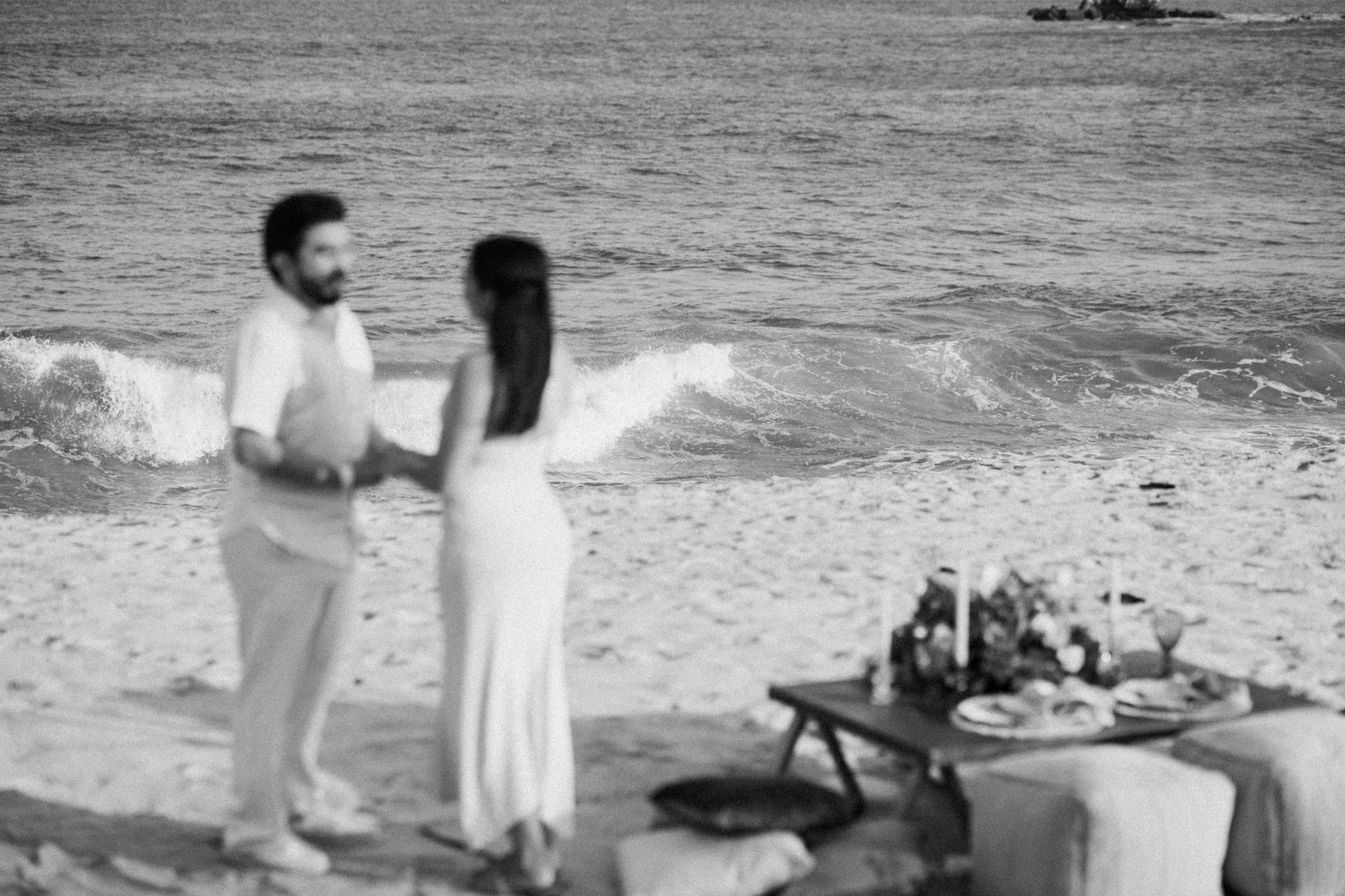 A romantic black and white beach scene with a couple dancing next to a picnic setup on the sand.