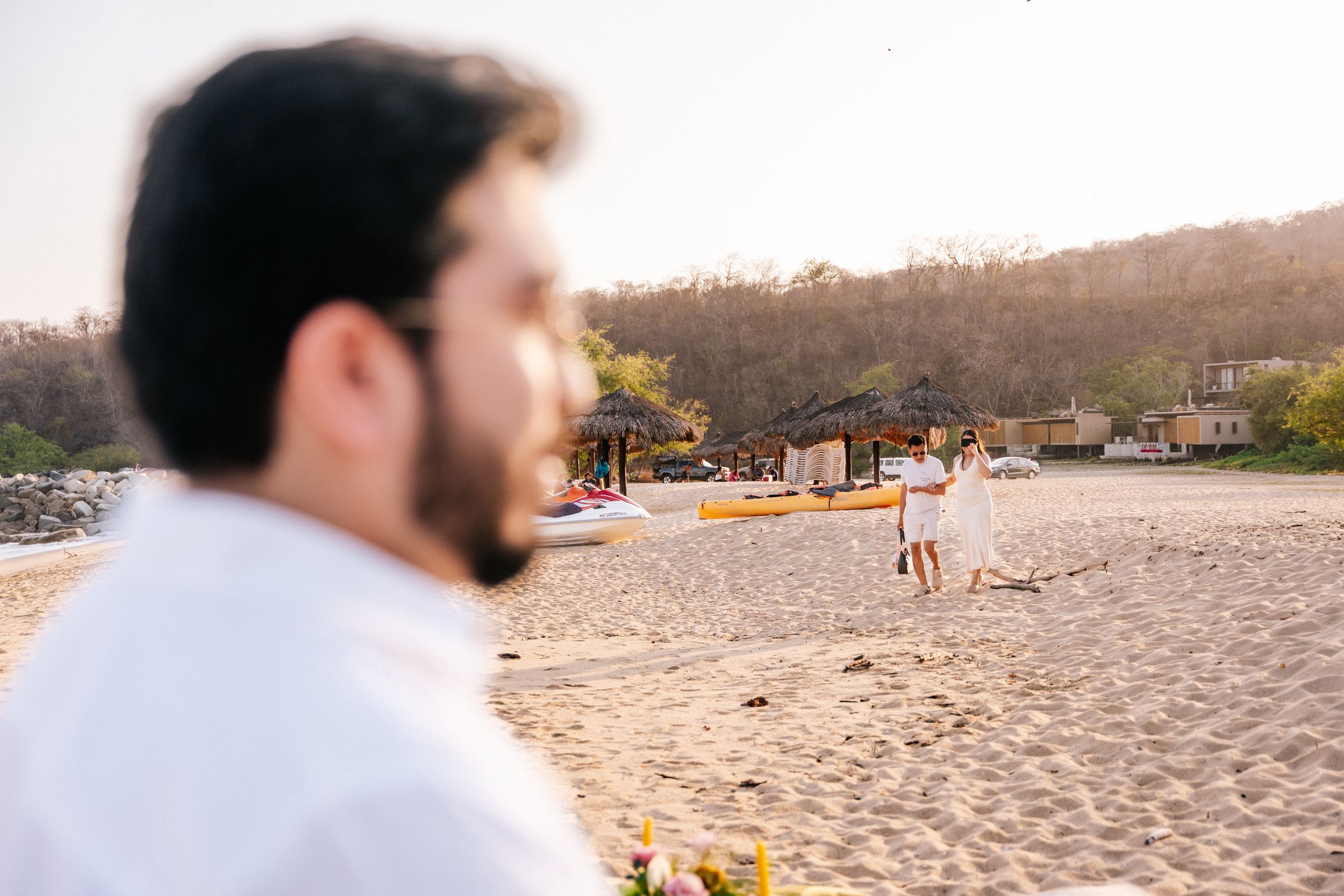 Blurred figure in white shirt looks out over a sandy beach at sunset with mountains in the background.