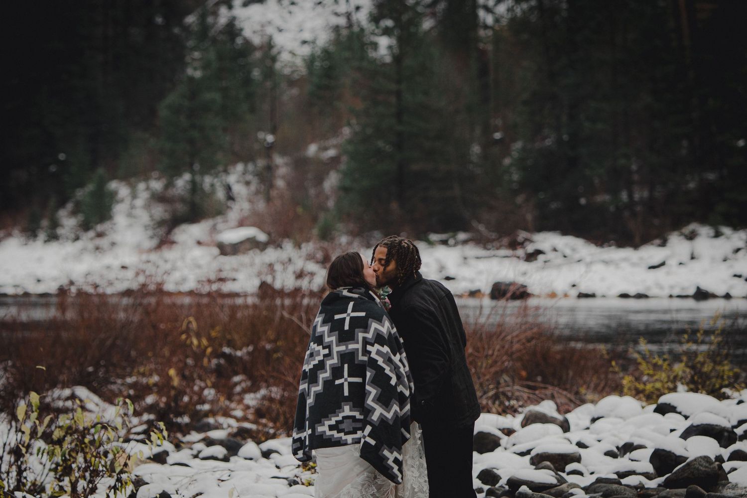A couple poses for wedding photos against a dramatic mountain backdrop with evergreen trees and a scenic lake.