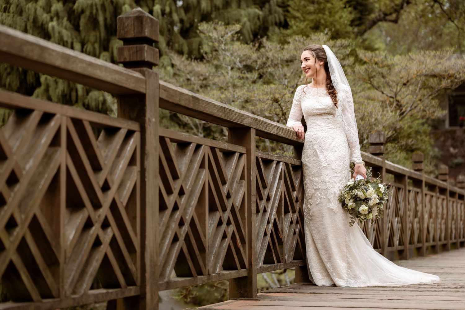 Bride in lace gown poses on rustic wooden bridge holding bouquet.
