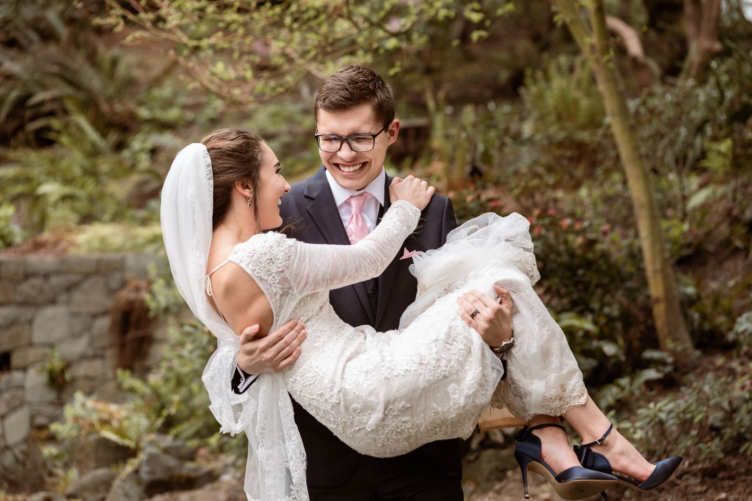 Groom carries bride in lace dress through a garden setting.
